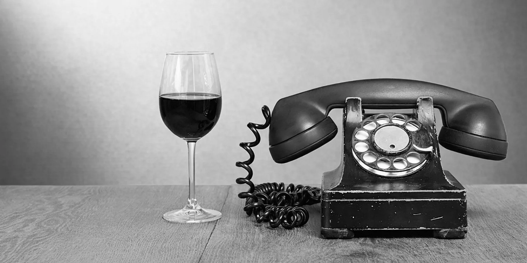 Black rotary phone with coiled cord next to a wine glass filled with red wine on a wooden surface