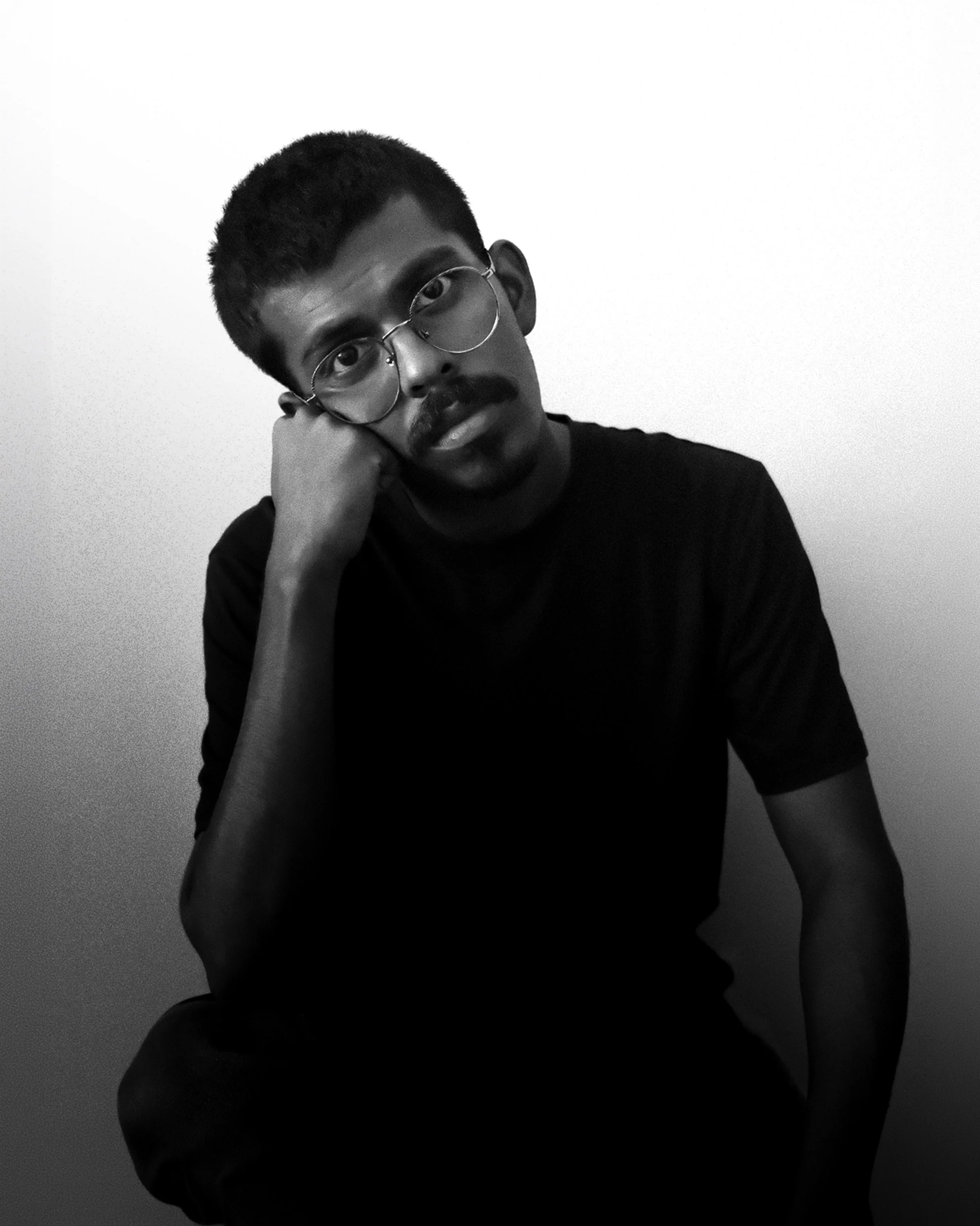 A black and white portrait of a young man with glasses, a mustache, and short hair, resting his face on his hand, sitting against a plain wall.