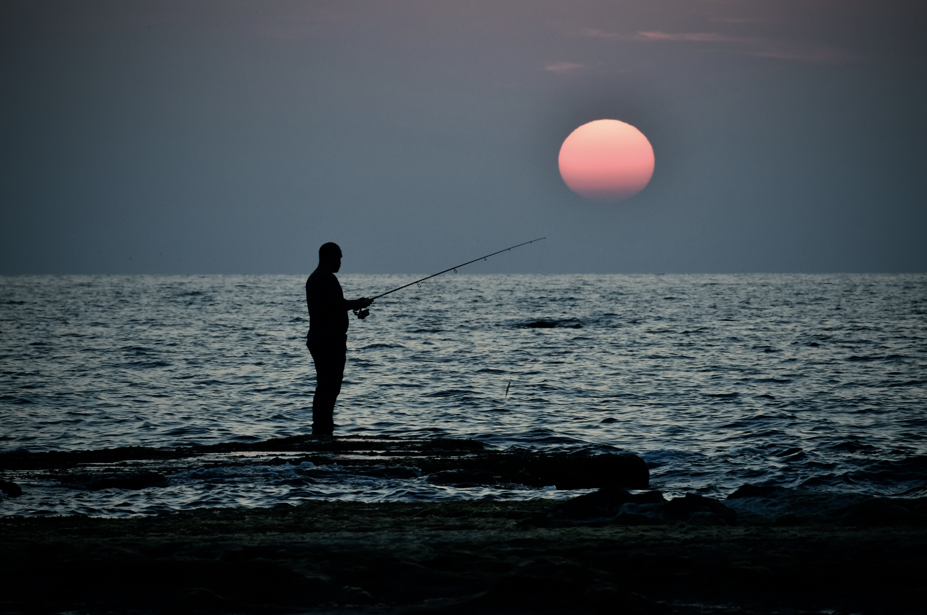 A person fishing at the shoreline during sunset with the sun setting on the horizon over the ocean.