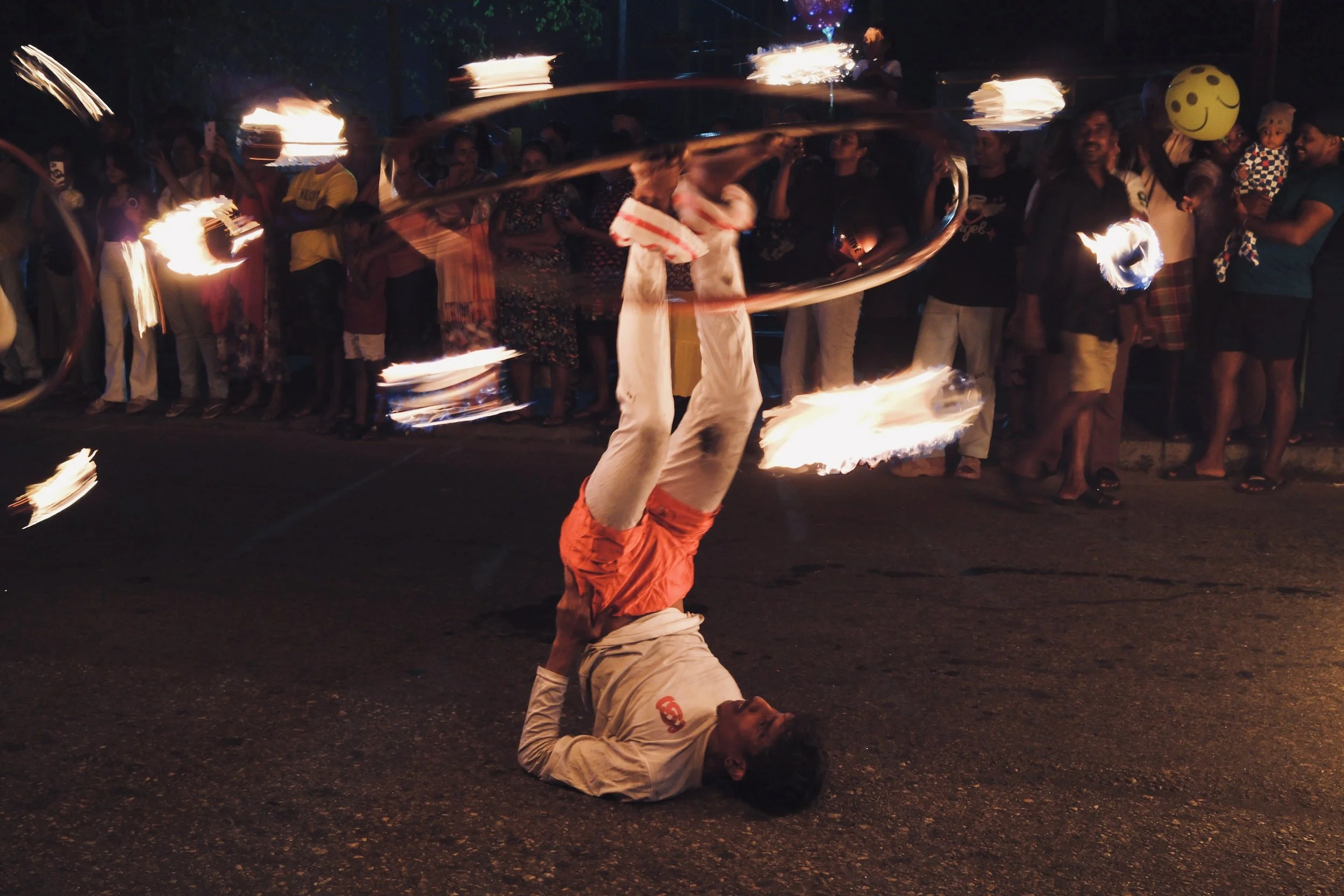 A person performing a fire dance act on the street at night, with fire poi spinning around them while they are lying upside down on the ground, surrounded by a crowd of onlookers.