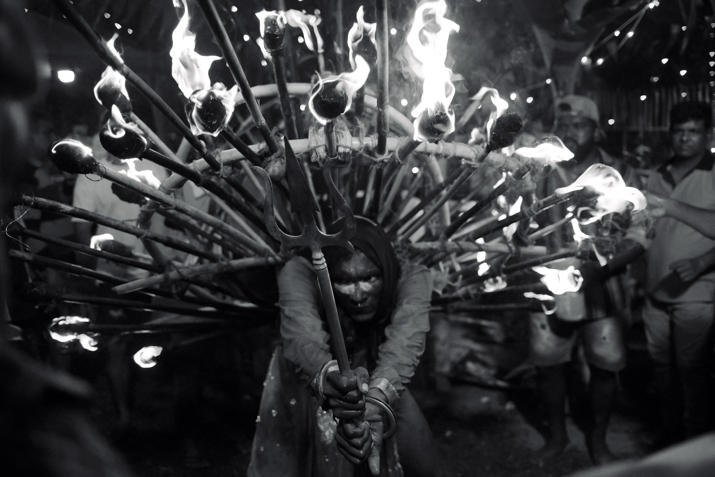 A person dressed in costume with multiple flaming torches arranged like a headdress, holding a staff with a trident at the end, during a performance at night, with onlookers in the background.