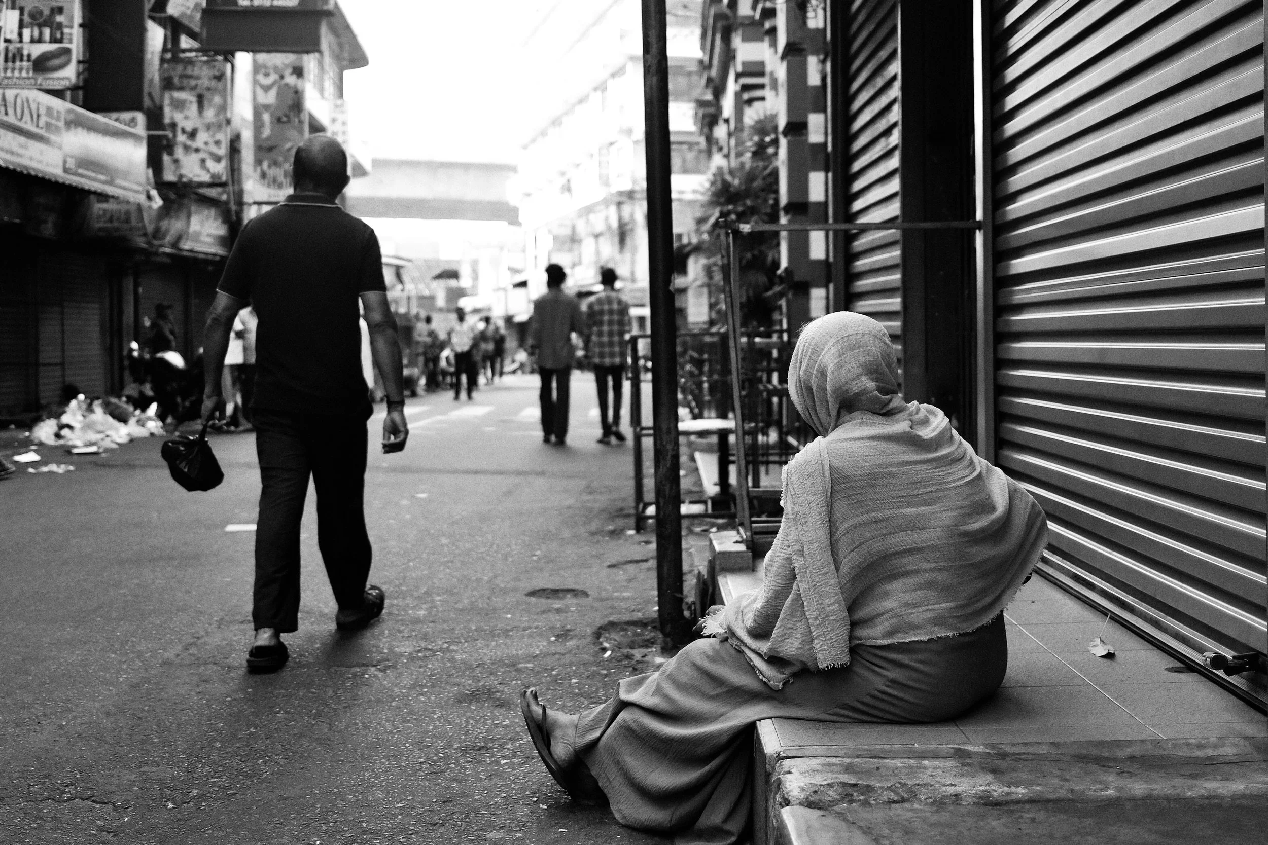 A woman wearing a headscarf sits on a sidewalk near a closed shop shutter while pedestrians walk along the street in an urban area.
