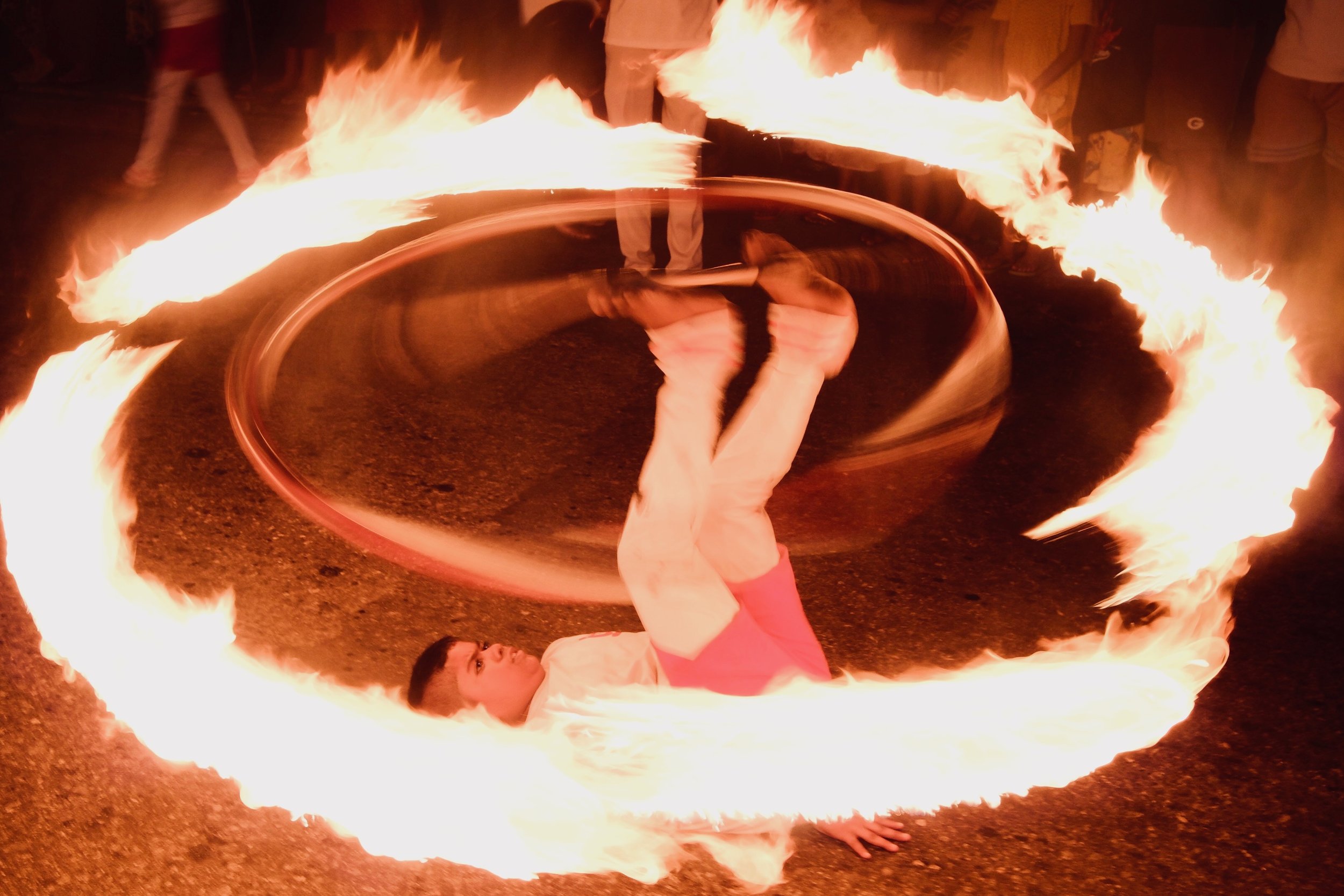 A person performing a fire spin trick with a fire hoop on the ground, creating a fiery ring, surrounded by onlookers.