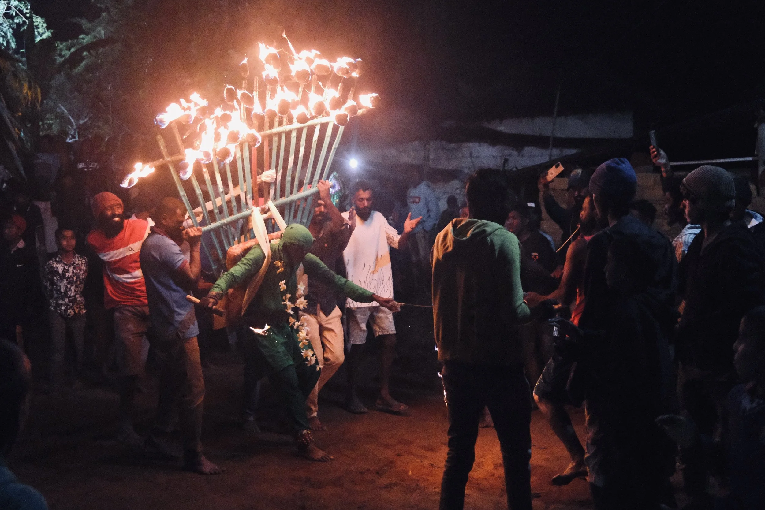 Group of people gathered outdoors at night, participating in a traditional fire ritual, with some dancing or performing around large flaming structure.