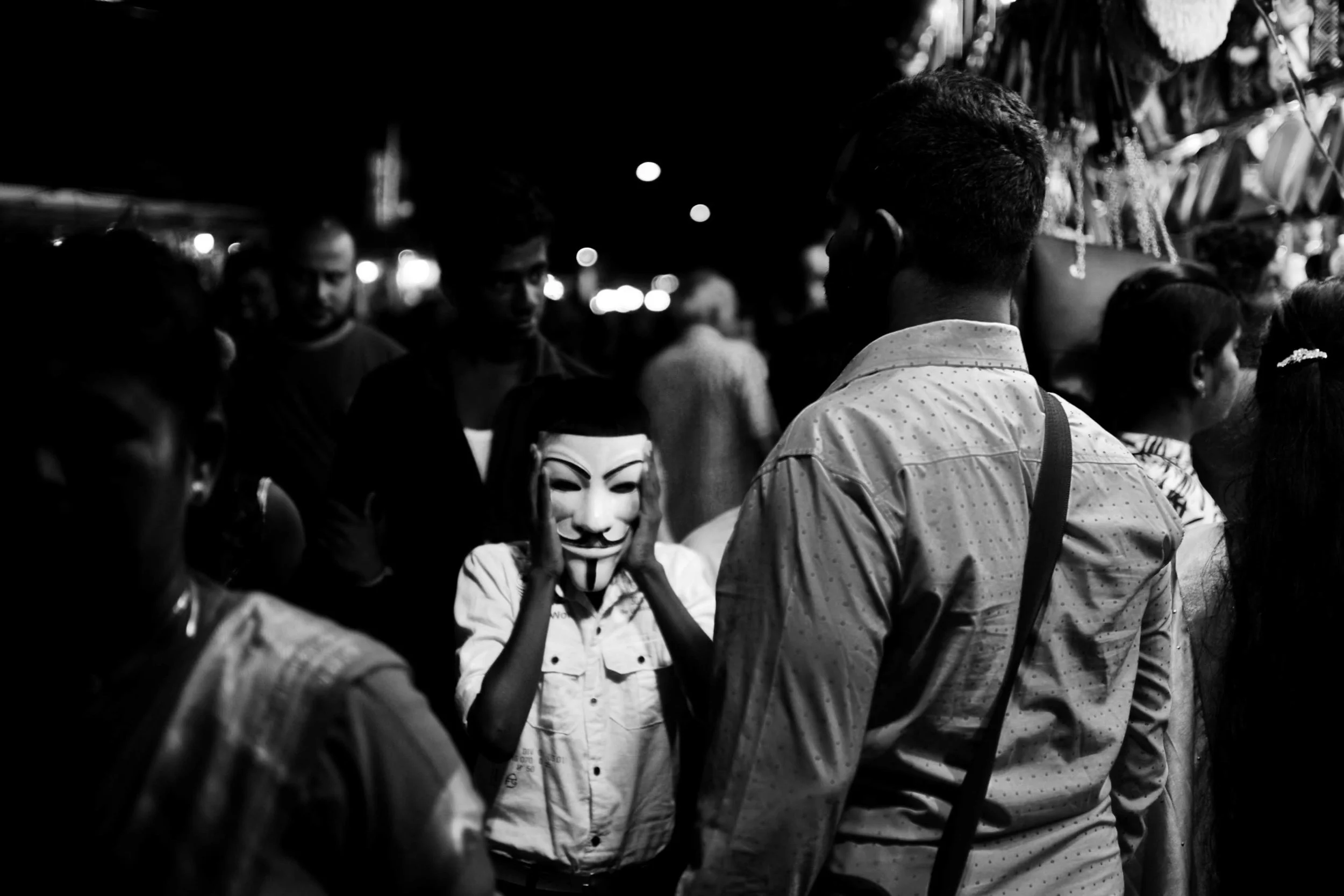 A person wearing a Guy Fawkes mask, touching their face, surrounded by other people at night.