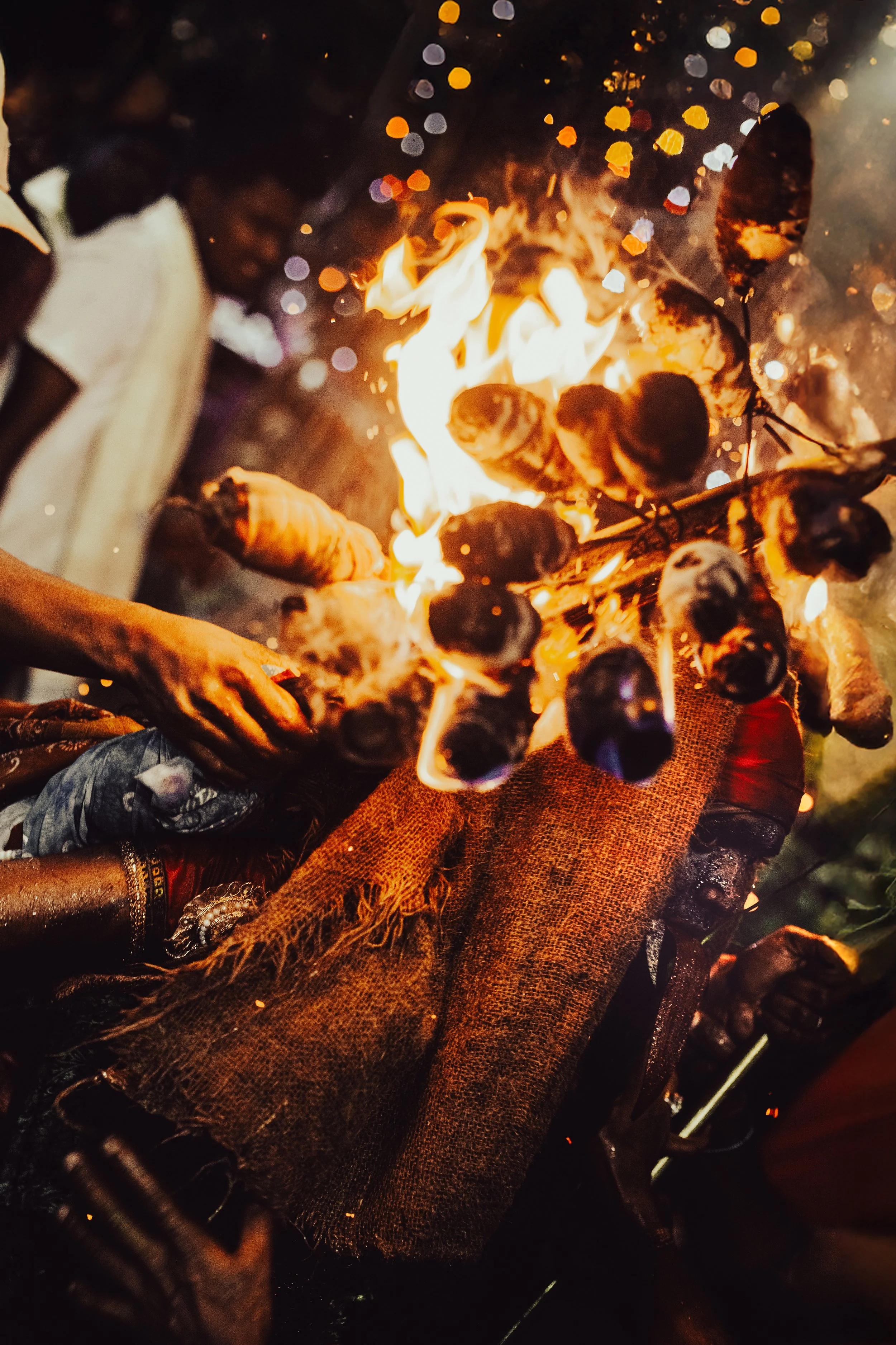 People gather around a bonfire with burning logs, at night, with colorful bokeh lights in the background.
