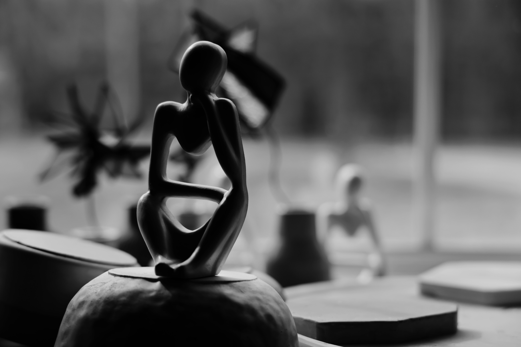 Black and white sculpture of a seated abstract human figure with one hand on its head, placed on a rounded base, with blurred objects and another sculpture in the background.