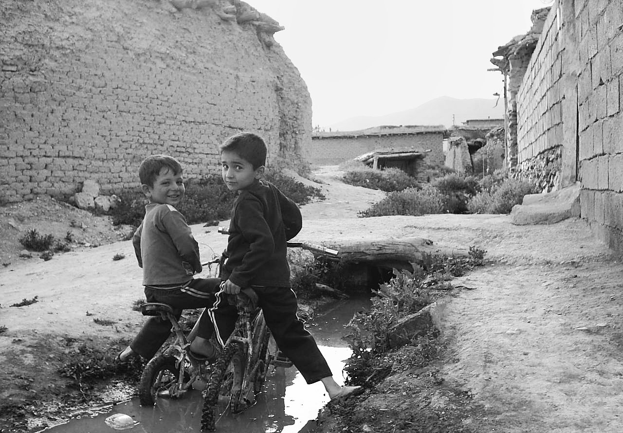 Two boys playing with a bicycle in a rural, muddy street with old brick buildings and mountains in the background.