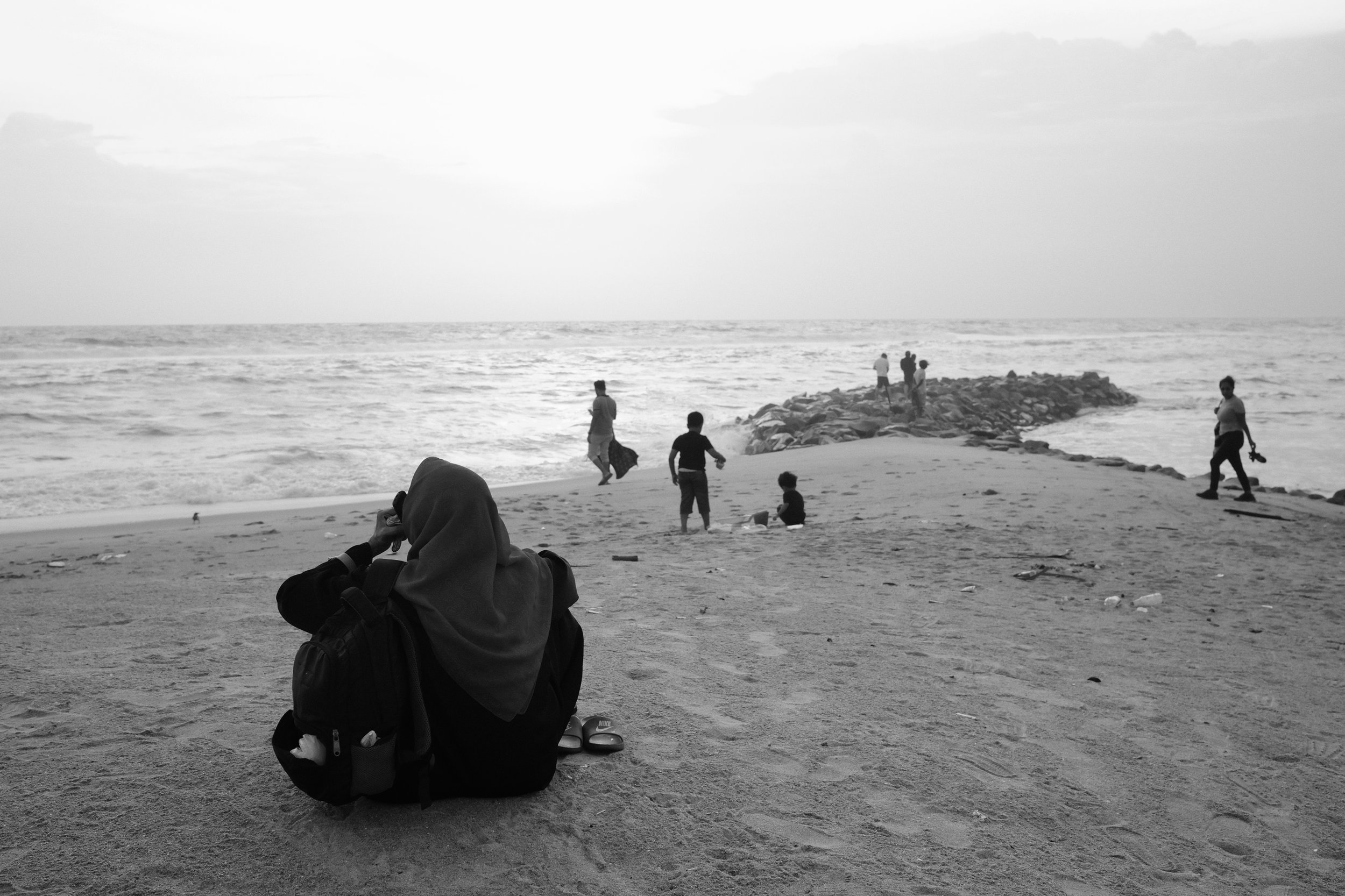A woman with a backpack and sandals sitting on the sandy beach, looking at the ocean, with a group of children and adults walking and playing near the ocean and on a rocky pier extending into the sea.