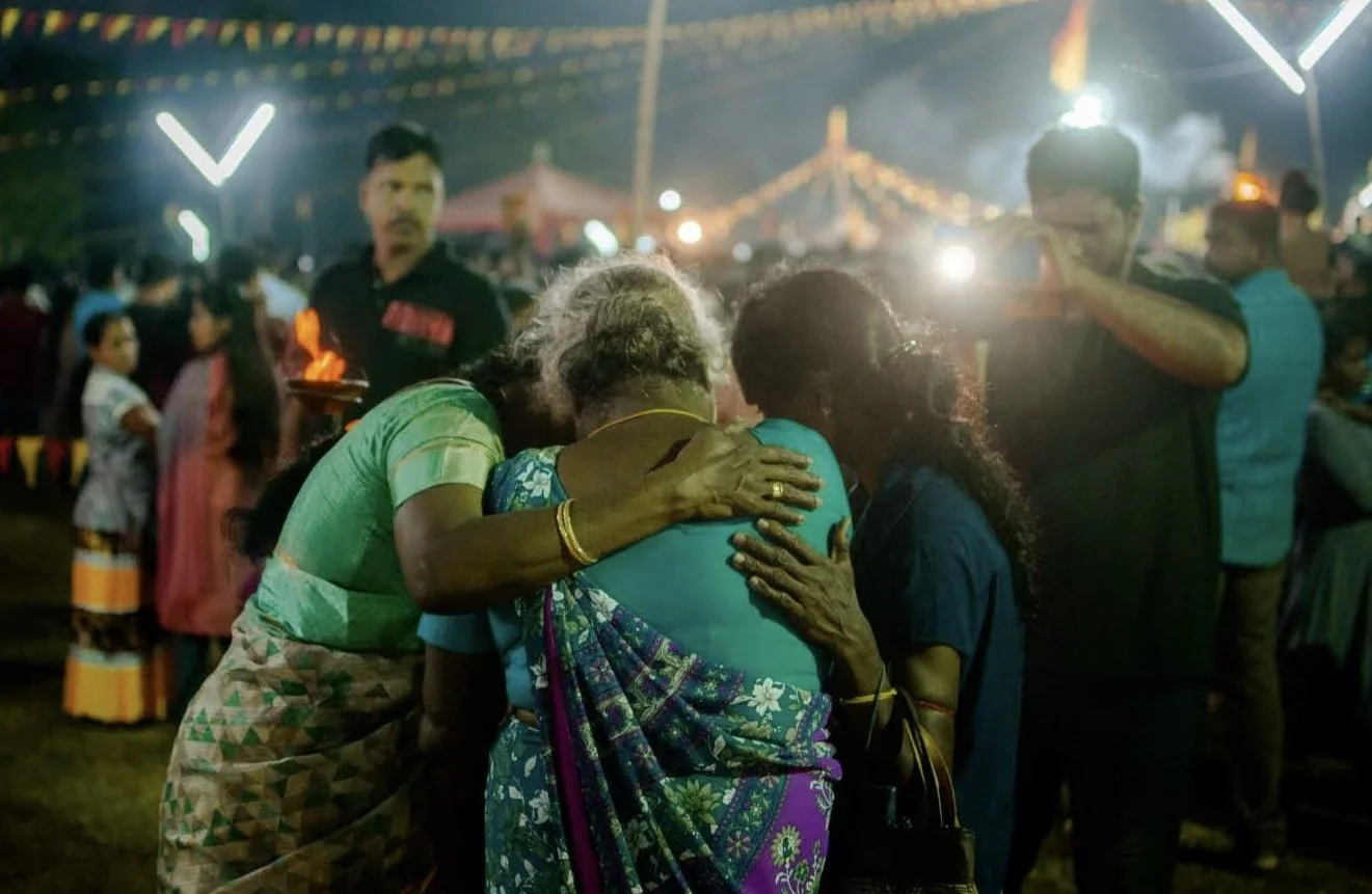 People at a nighttime festival or fair, with some emotional moments; a woman and two older women are hugging, and a man and woman are nearby, with festive lights and tents in the background.