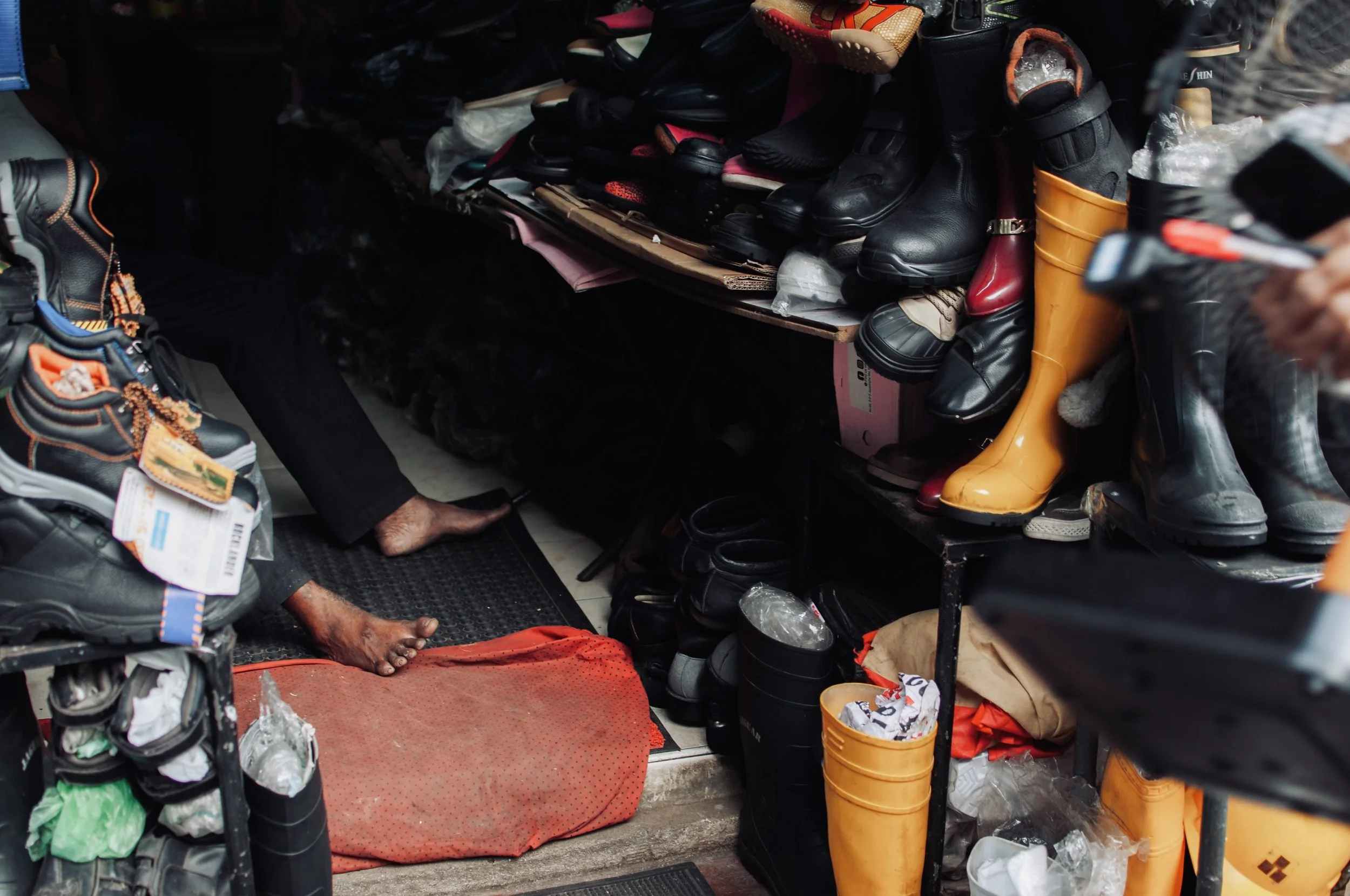 Inside a shoe store or repair shop, shelves filled with various shoes including boots and sneakers, with a person sitting on the floor, partially visible, barefoot with a dark complexion and wearing dark clothing.