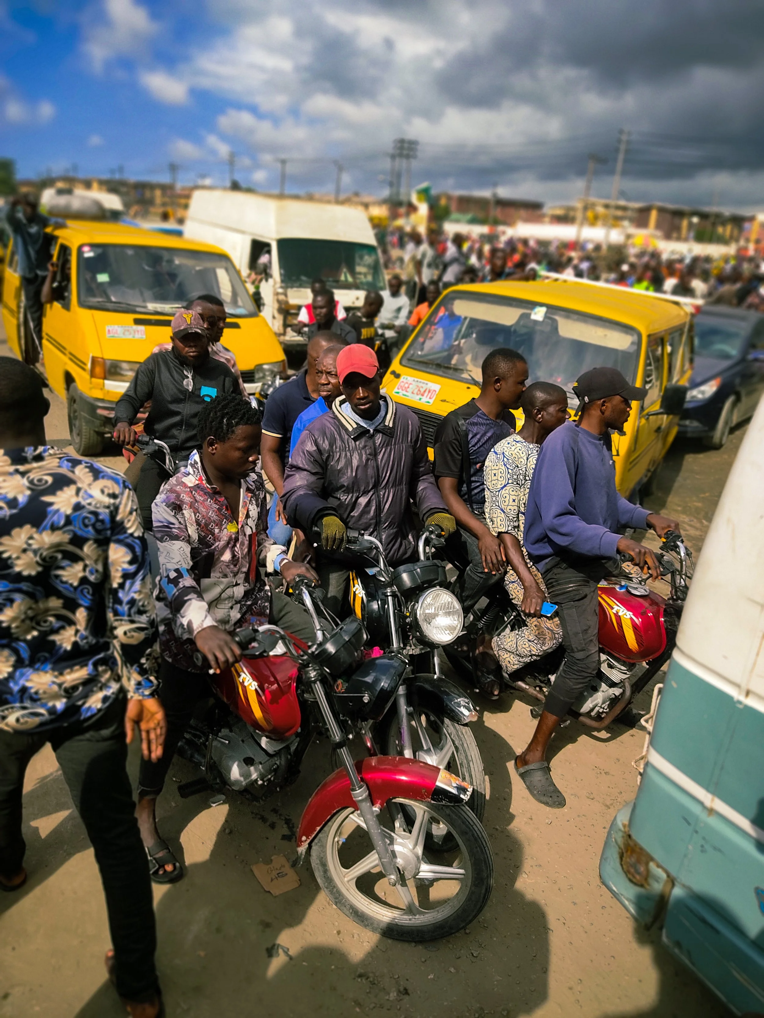 A crowded street scene with many people, motorcycles, and small yellow tuk-tuk vehicles. The sky is partly cloudy with dark clouds.