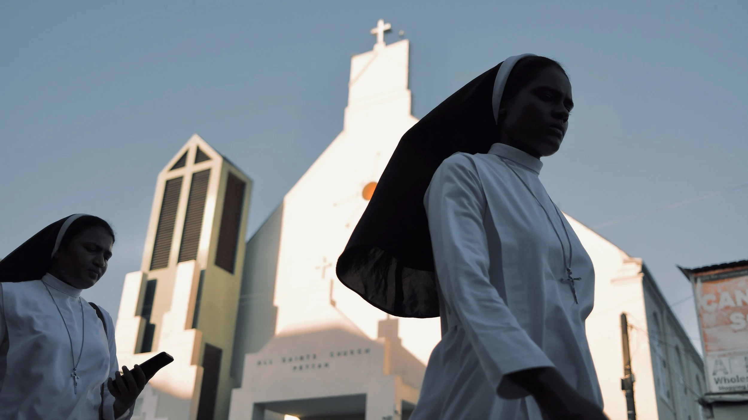 Two nuns walking on the street, one looking down at a phone, in front of a church with a cross on top.