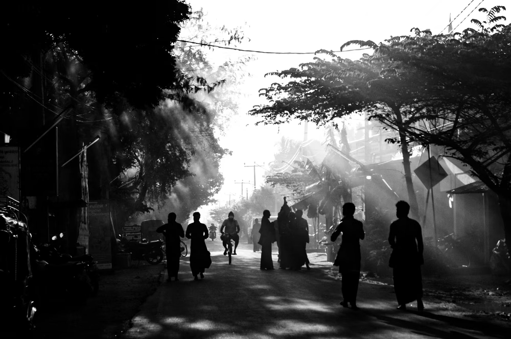 Silhouettes of people walking on a street with damaged buildings and trees, black and white photo.