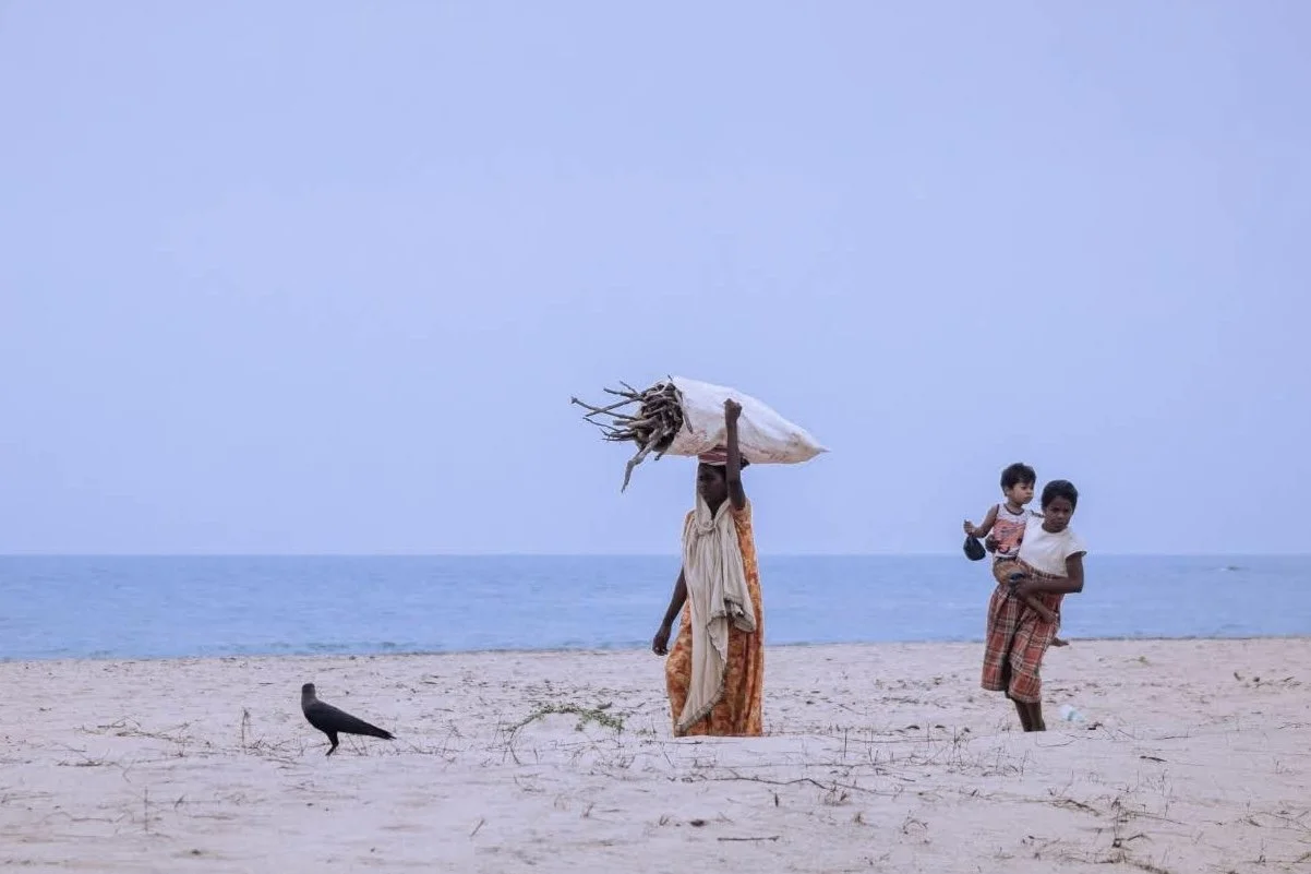 People walking on a sandy beach near the ocean, with one person carrying a bundle of dried sticks or roots on their head, and a woman holding a child.