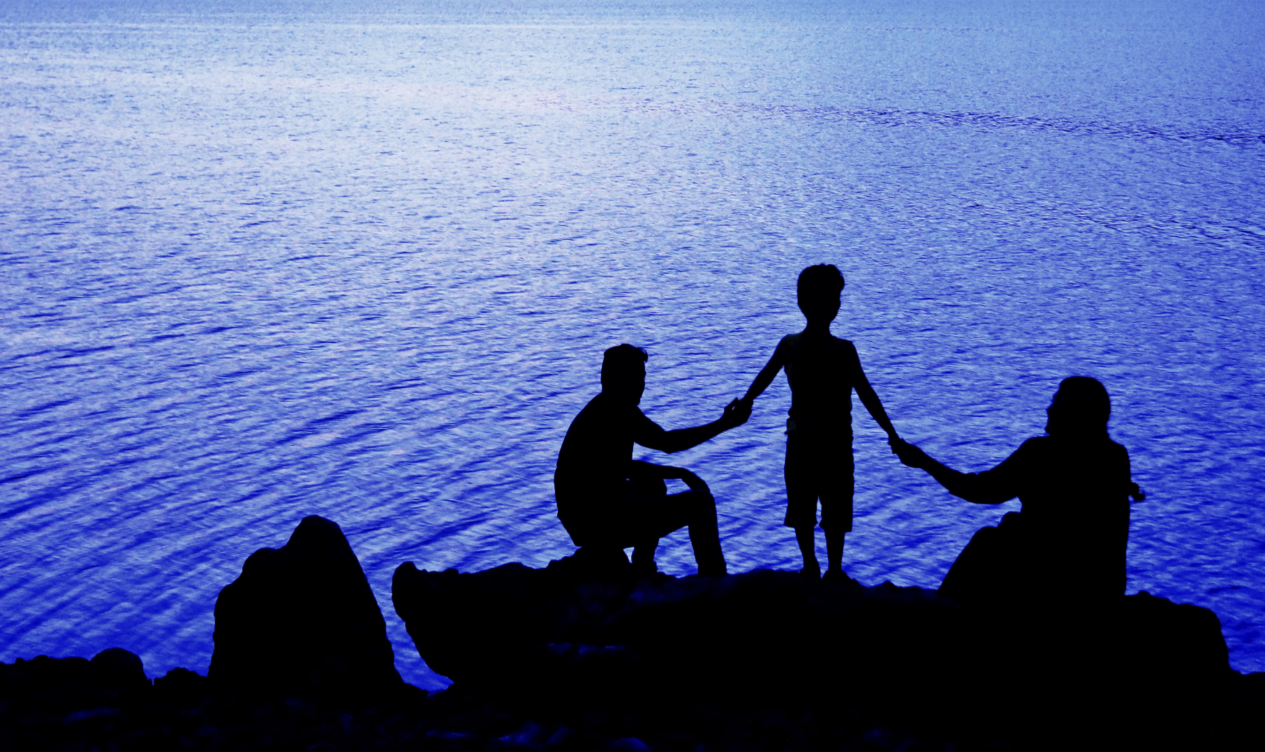 Silhouettes of three children on rocks by water at dusk, two holding hands with a third child standing between them, overlooking a calm blue body of water.