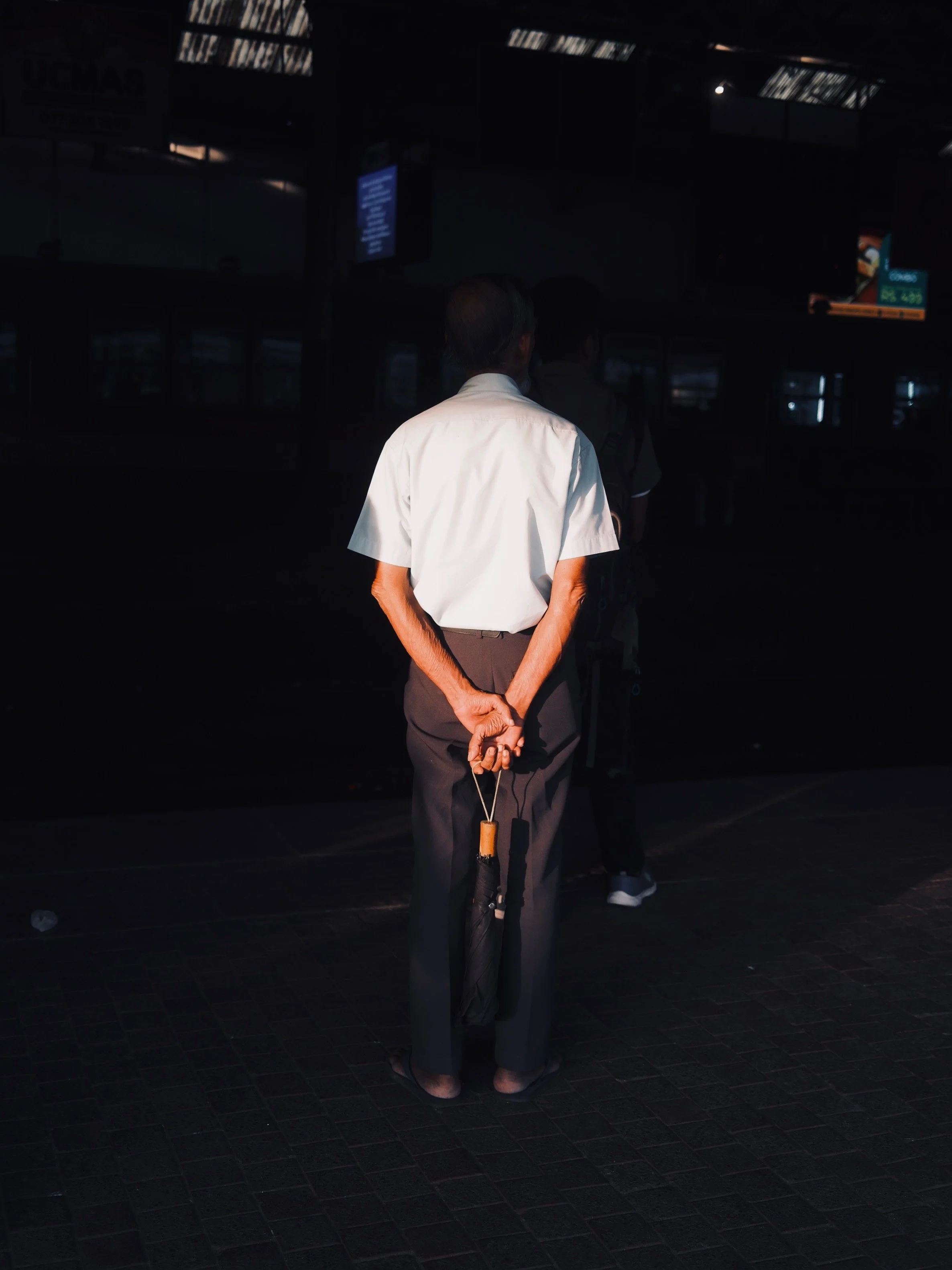 Man standing with hands clasped behind back, holding an umbrella, at a train station or bus terminal, with dark background and some people walking in front.