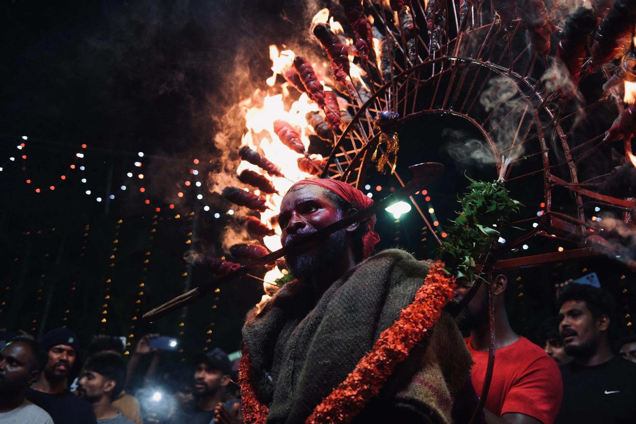 A performer with dark makeup and a red headband, carrying a large flames-lit decorative structure on his back, participating in a traditional festival surrounded by a crowd at night.
