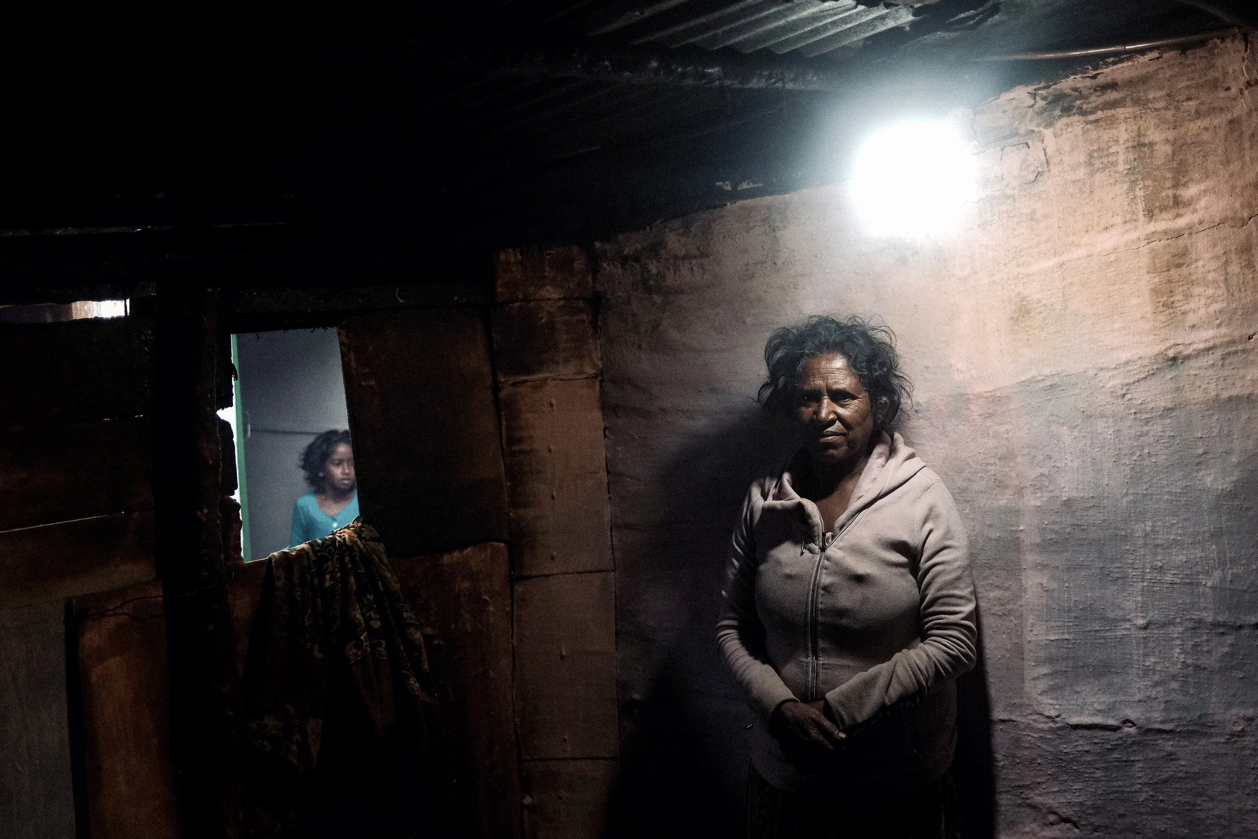 An elderly woman with curly hair stands inside a small, dimly lit room with wooden walls. A bright light is visible from the ceiling, and a young girl is seen in a doorway behind her.