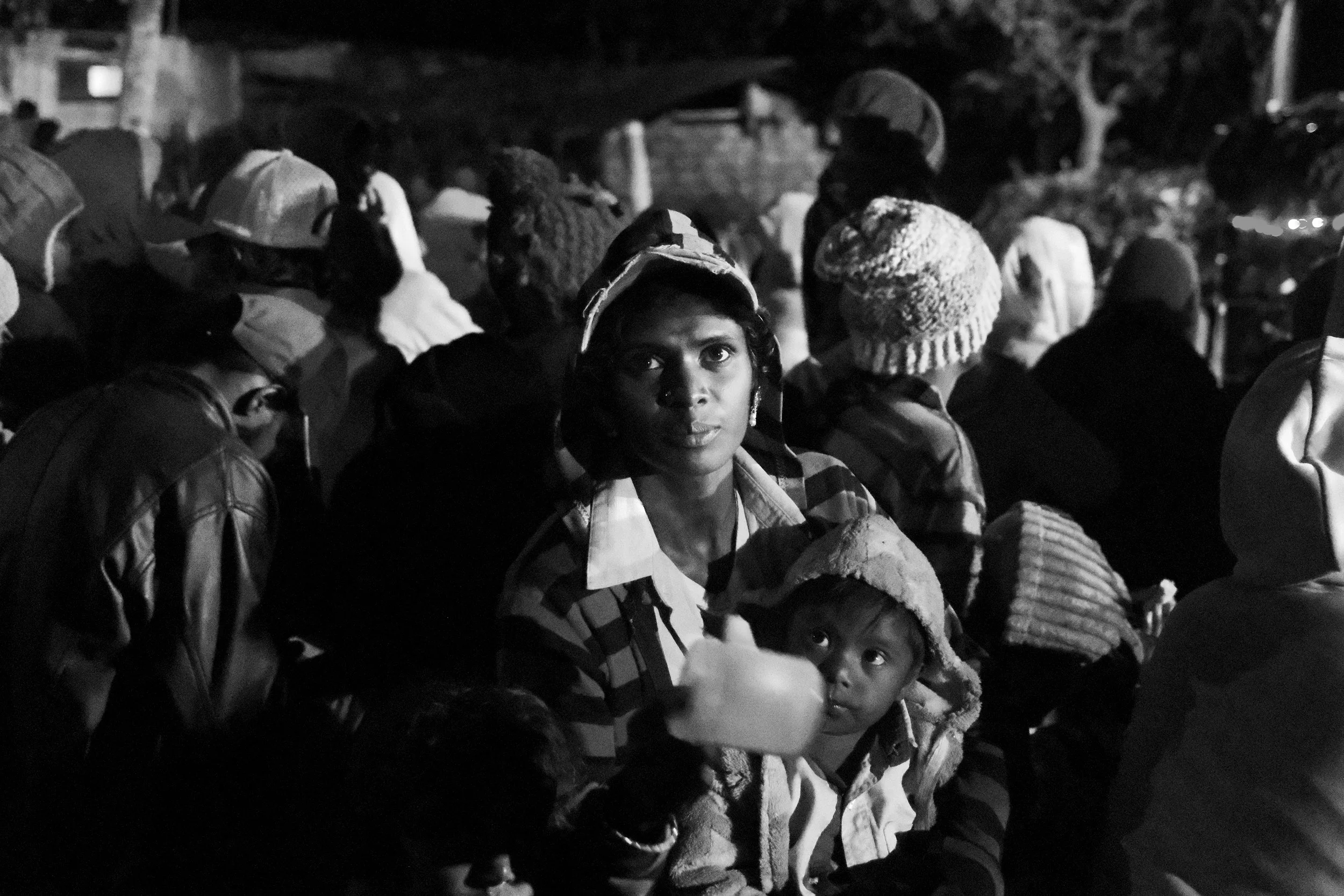 A woman and a young girl sitting among a crowd of people at night, some wearing warm clothing and hats, with trees and a bridge in the background.