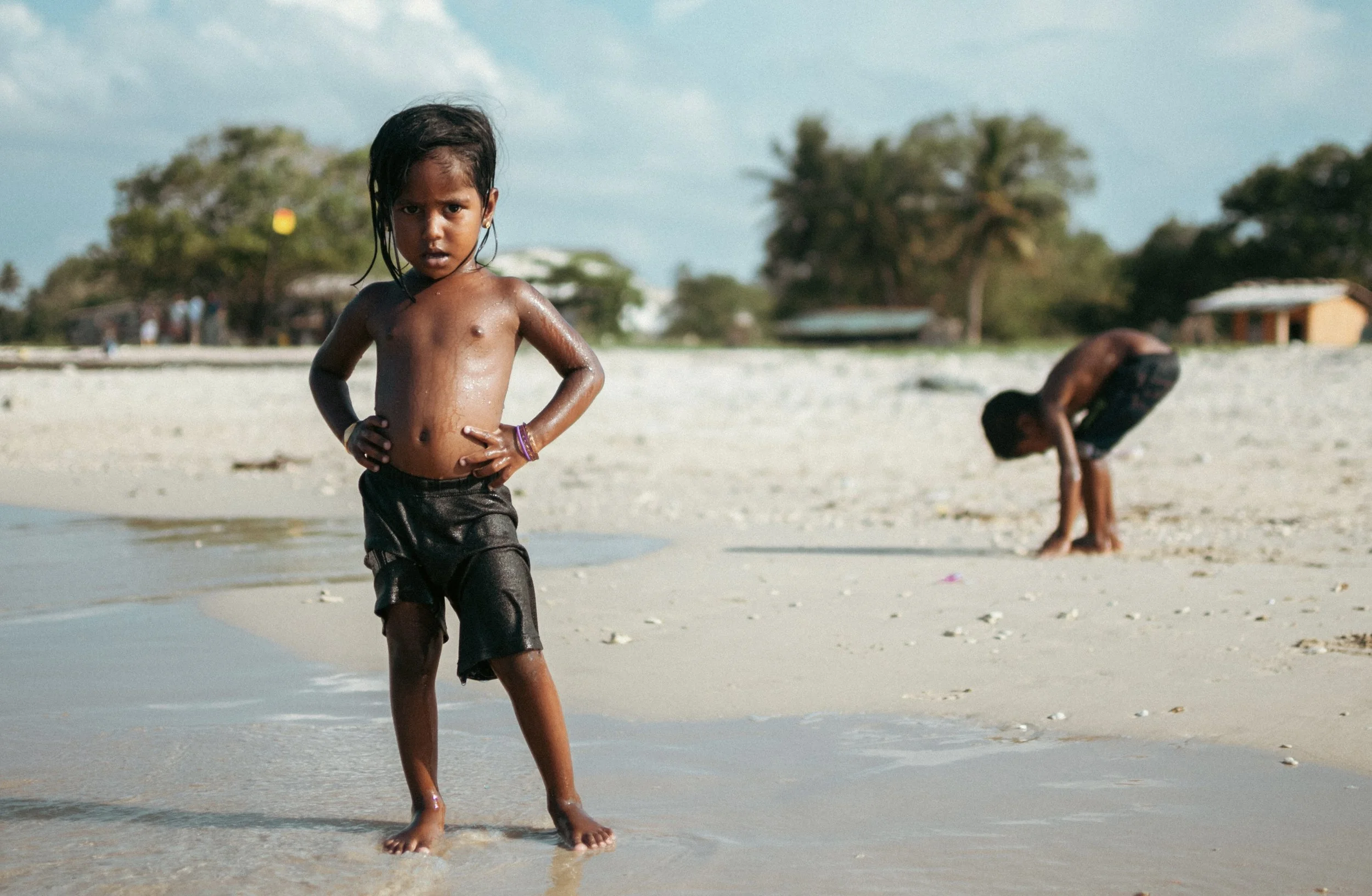 A young girl standing on the beach with hands on her hips, looking at the camera, with another child bending down near the water in the background.