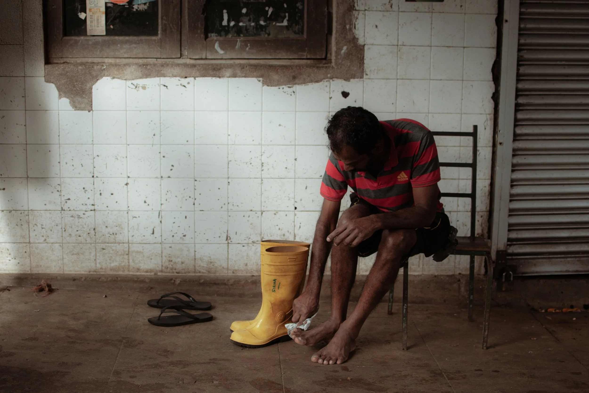 A man sitting on a chair on a dirty floor in front of a tiled wall, putting on a yellow rubber boot. A pair of sandals is on the floor nearby.