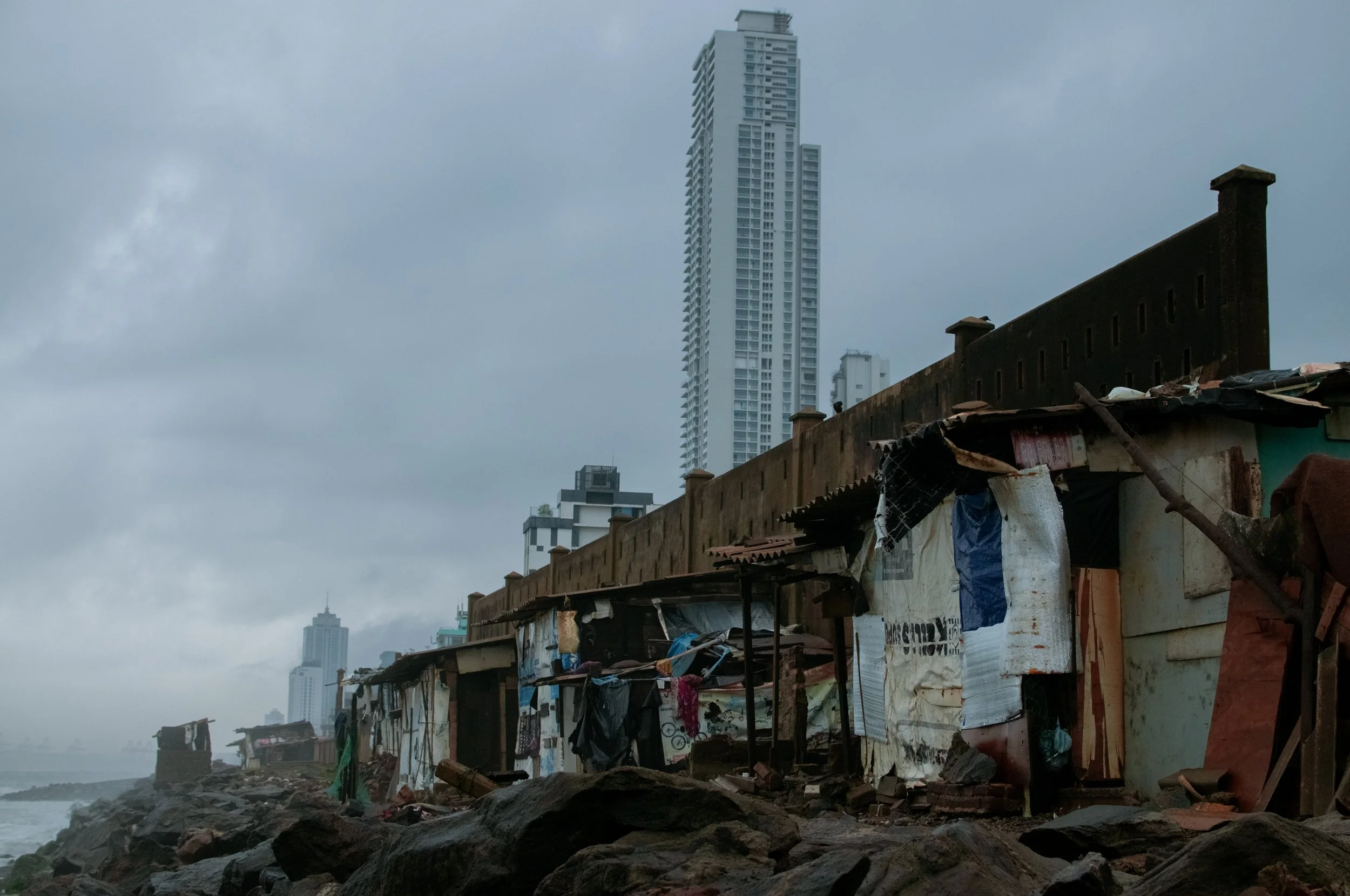 Makeshift shacks built from scraps of wood and fabric along a rocky shoreline with high rise buildings in the background under cloudy, overcast sky.