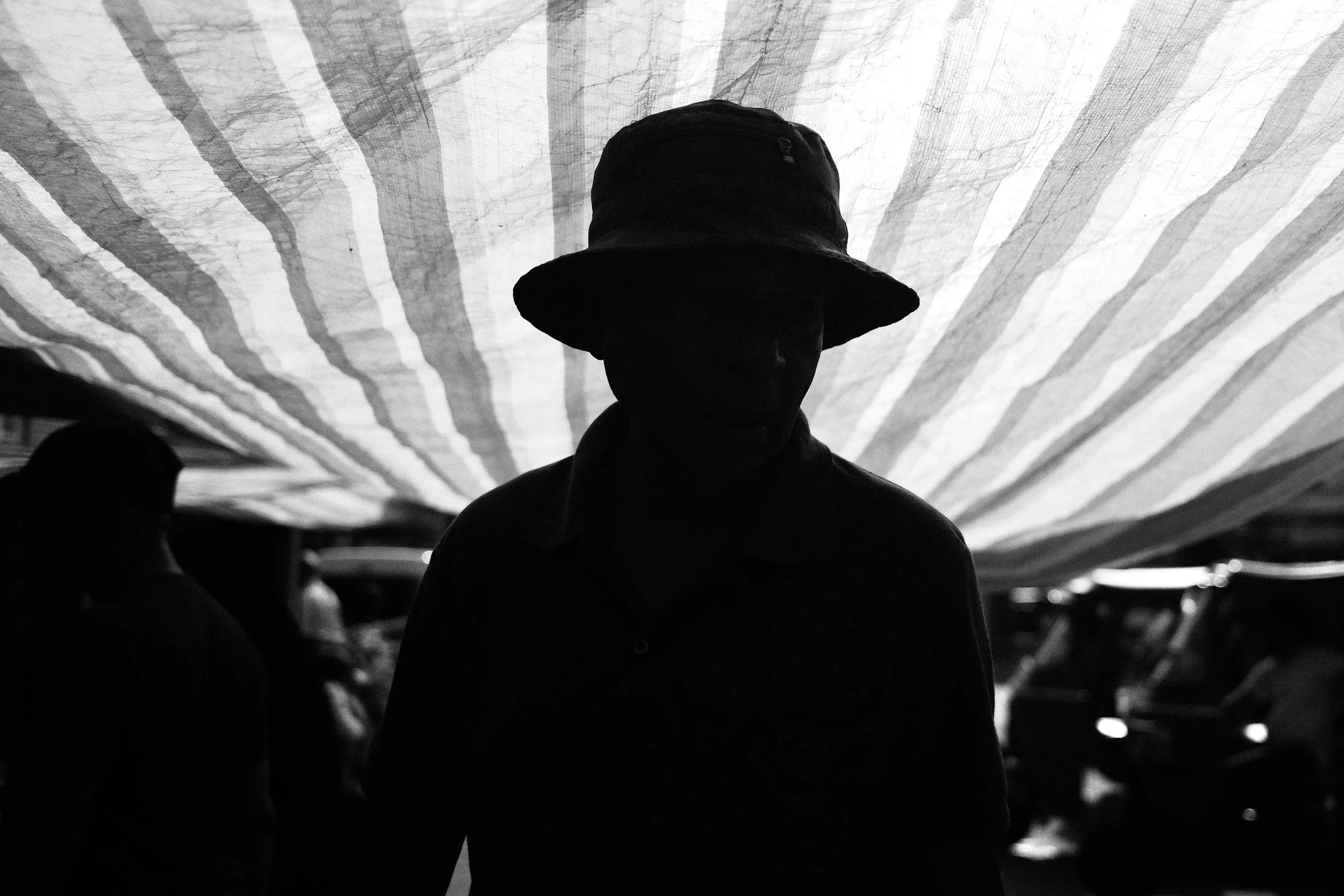 A person wearing a hat standing in front of a striped fabric canopy with other people and vehicles visible in the background.