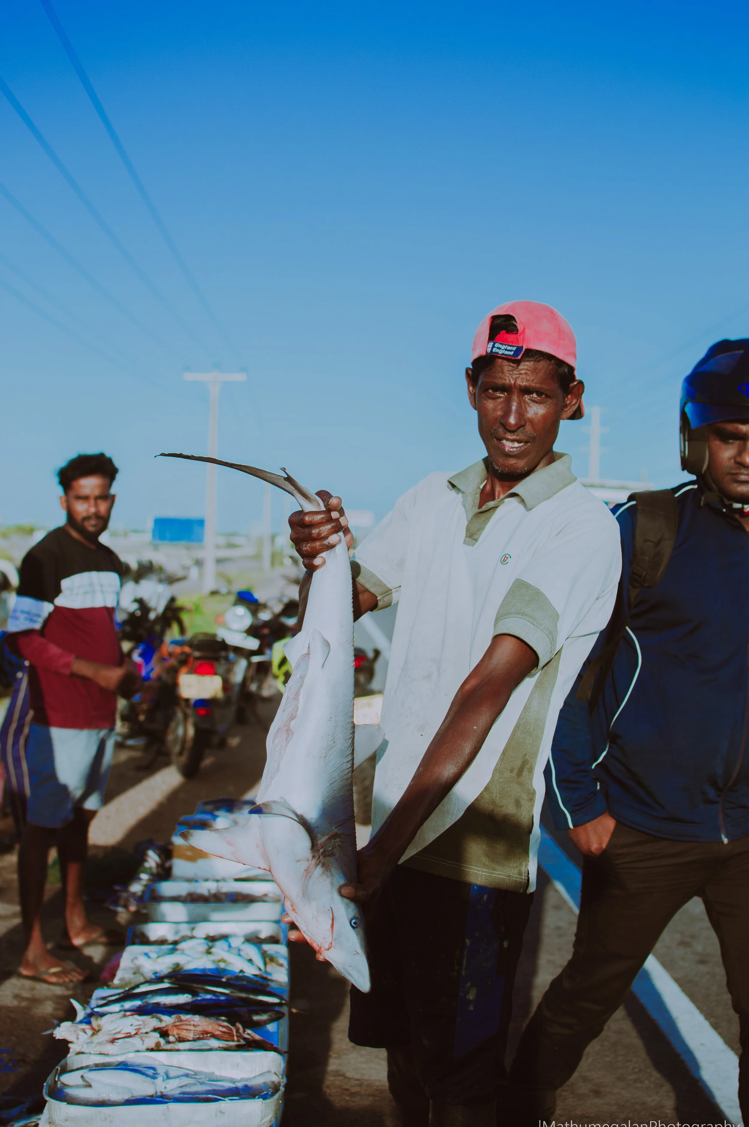 Man holding a large fish at a market, with other fish laid on a table and motorcycles in the background.