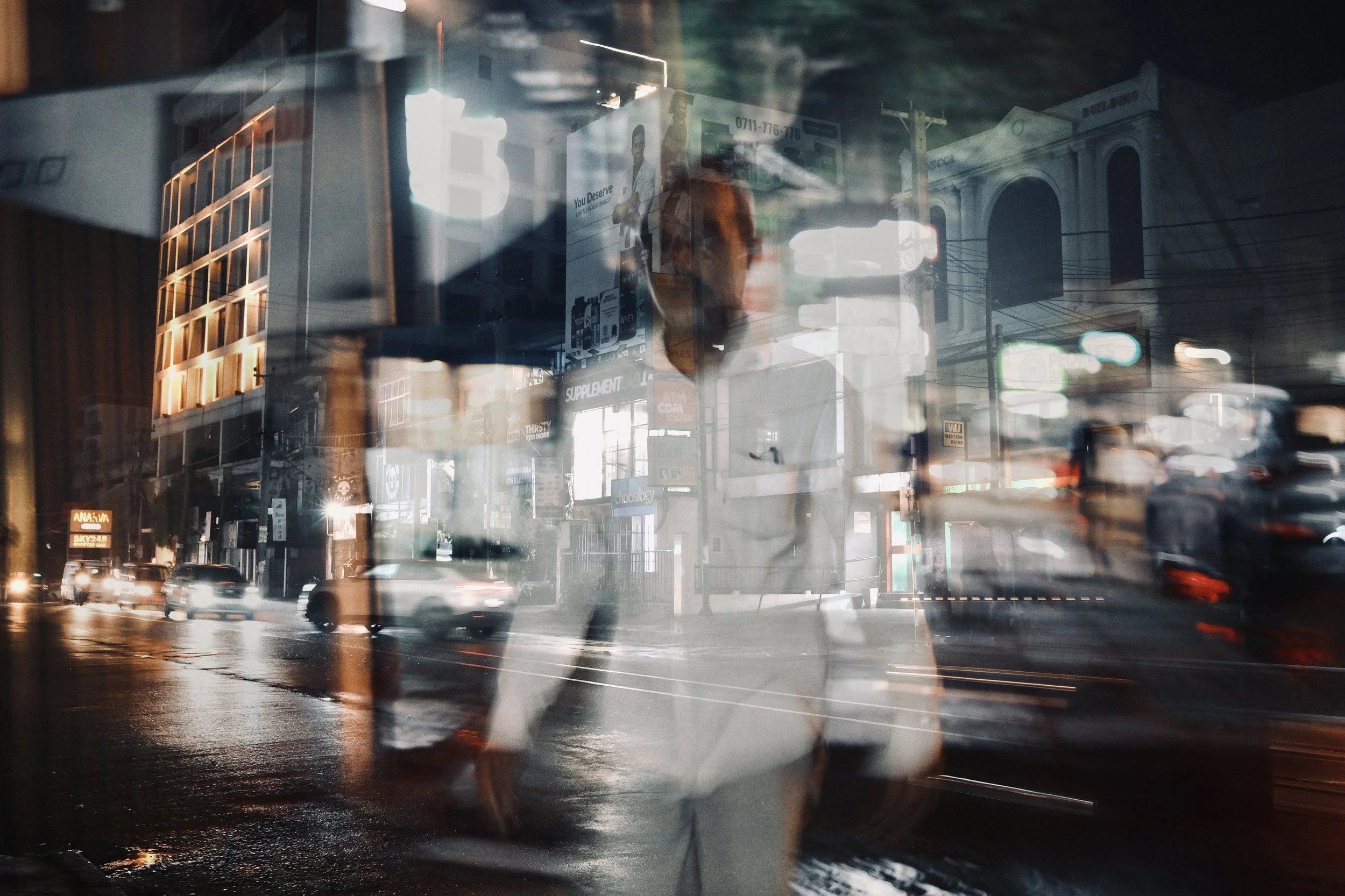Night city street scene with reflections of buildings, vehicles, and a person in the glass.