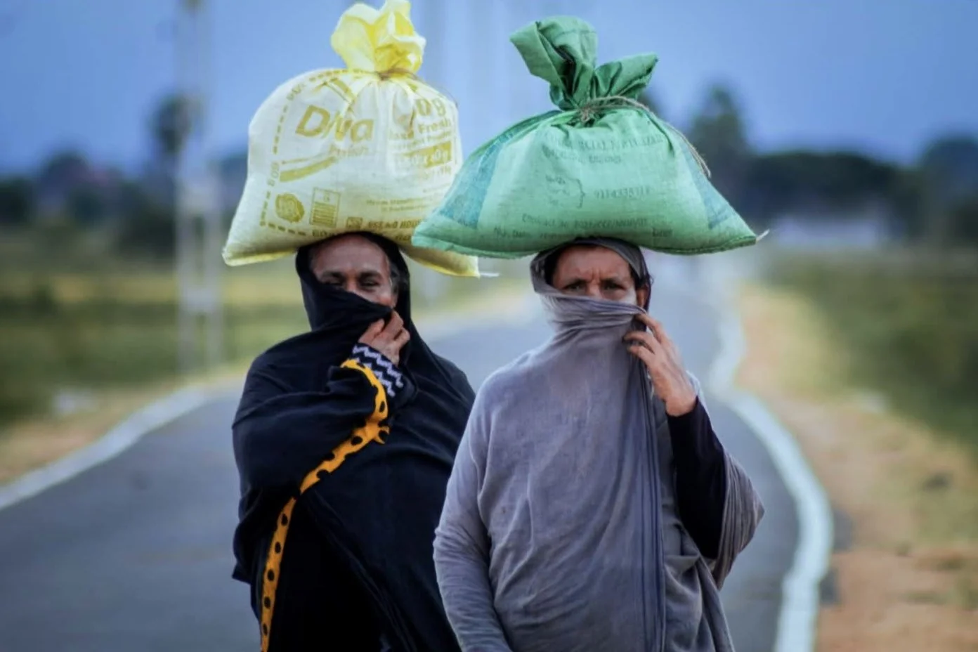 Two women walking on a rural road, carrying bags on their heads, with their faces partially covered by scarves.