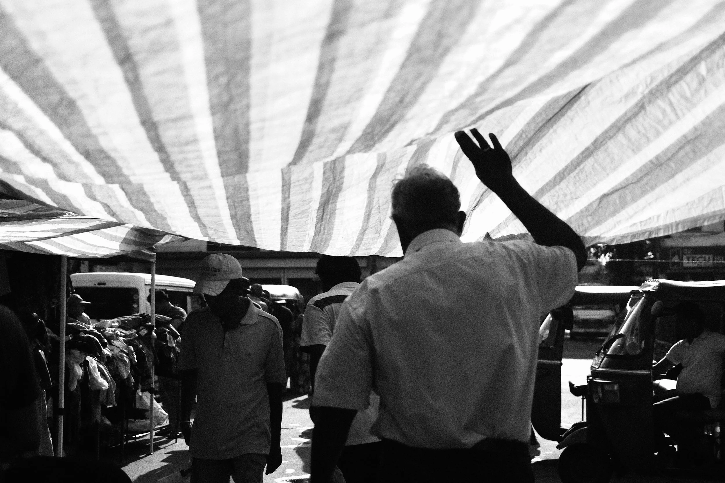 Black and white photo of a busy outdoor market scene with people walking and various stalls, including a person waving under a large striped canopy.