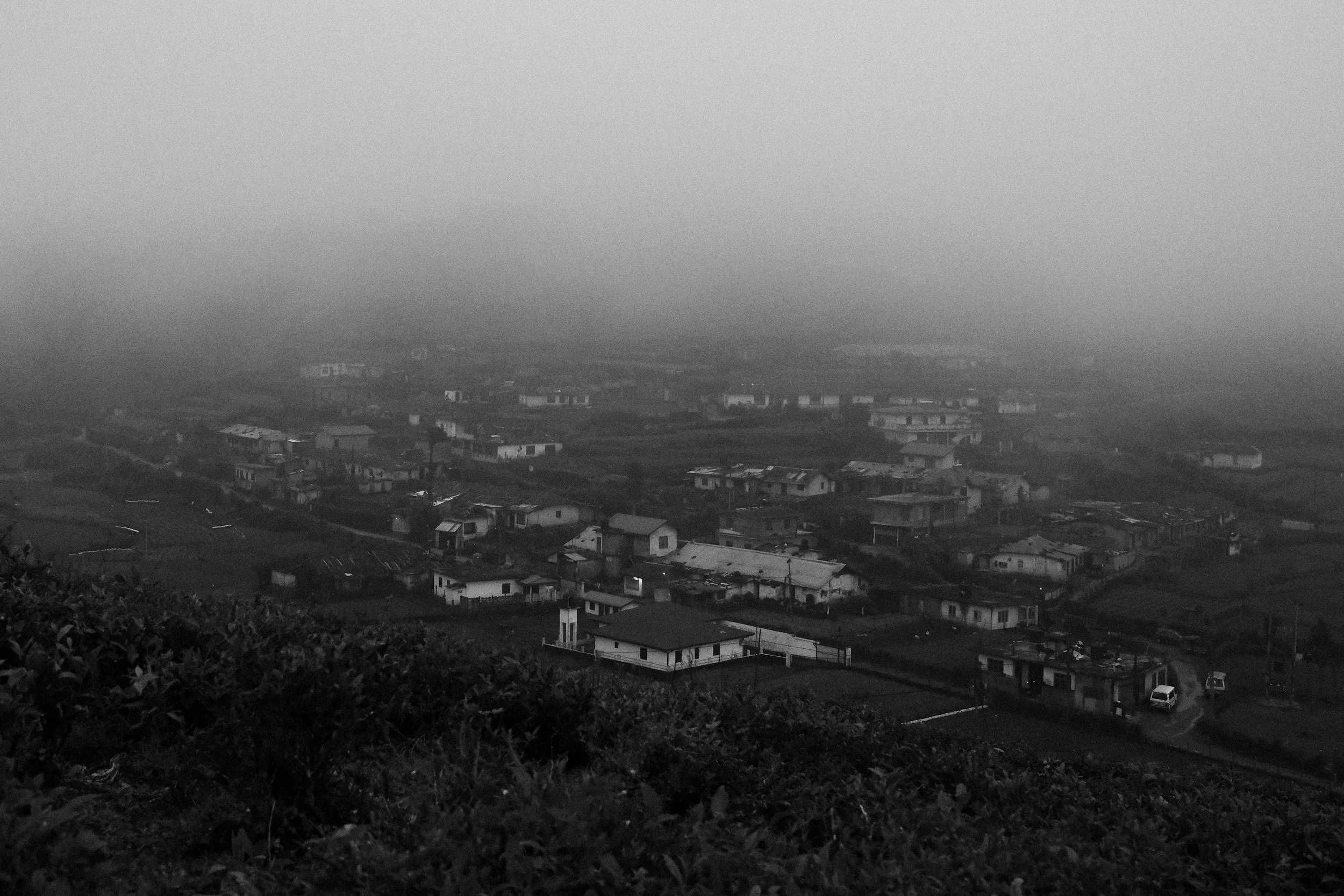 A black and white photo of a small town or neighborhood on a hillside with houses and roads, shrouded in fog or mist.