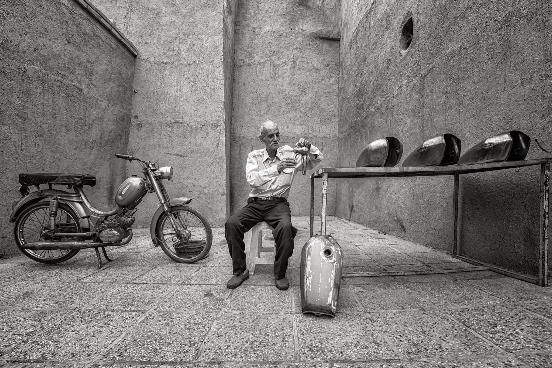 An elderly man sitting on a stool, decorating a cross with a string of beads, next to an old motorcycle and a table with five oil cans, in a narrow alley with textured walls.