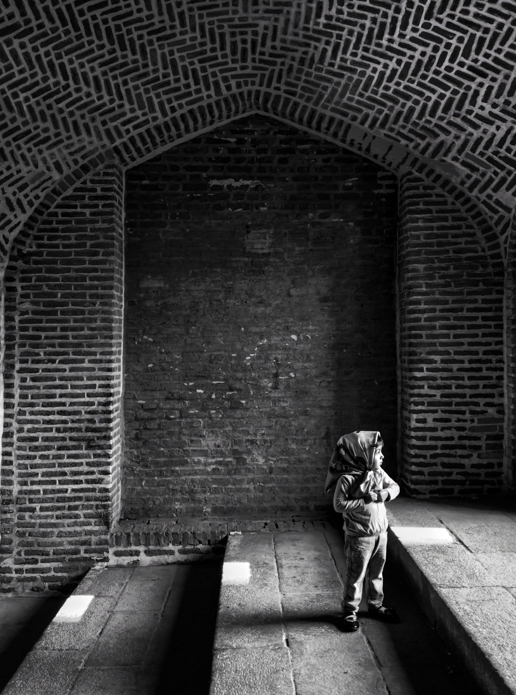 A young girl standing on a stone platform under a large brick archway, with shadows cast on the ground.