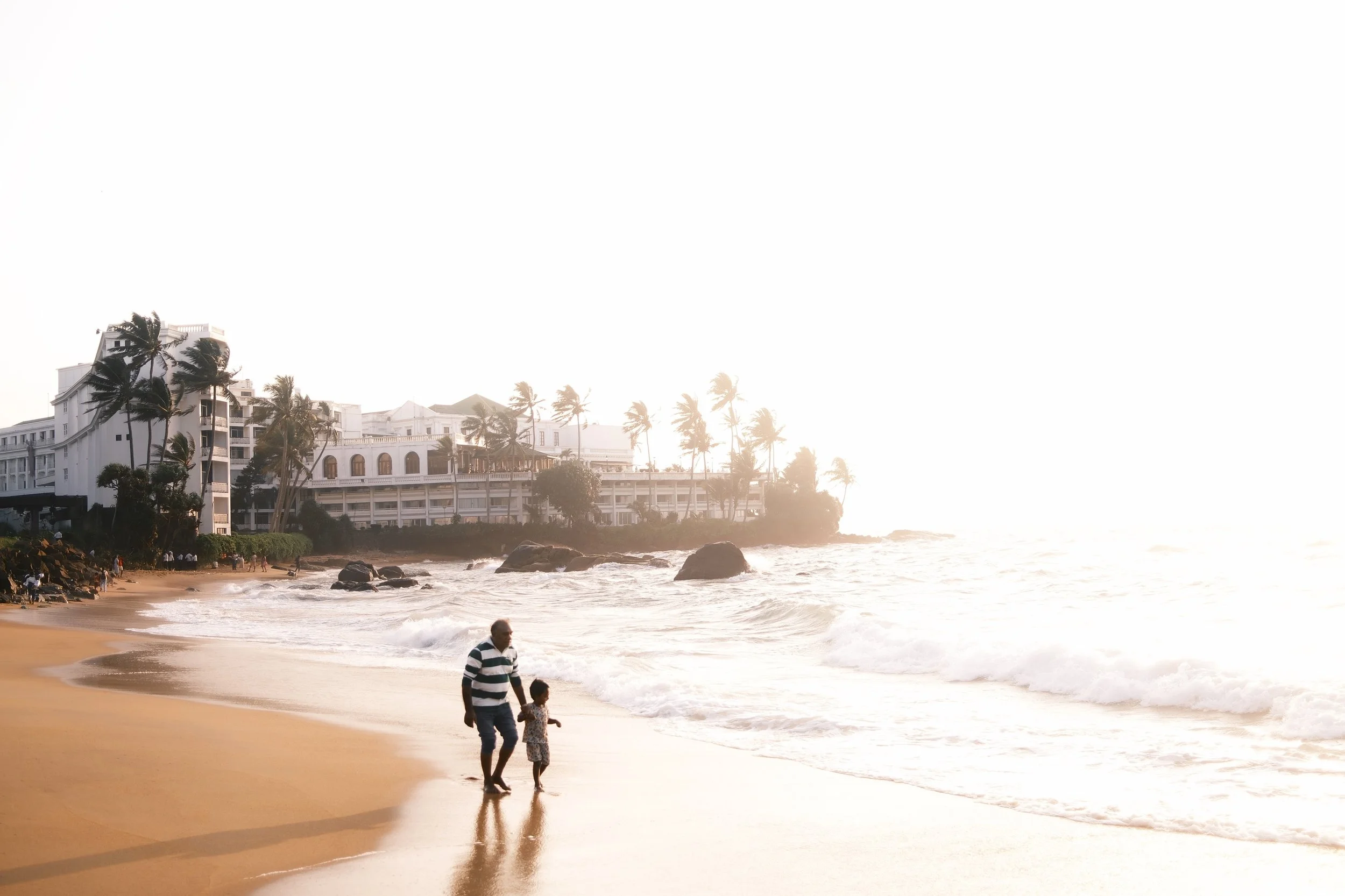 Father and child walking along the beach at sunset, with a large white building and palm trees in the background.