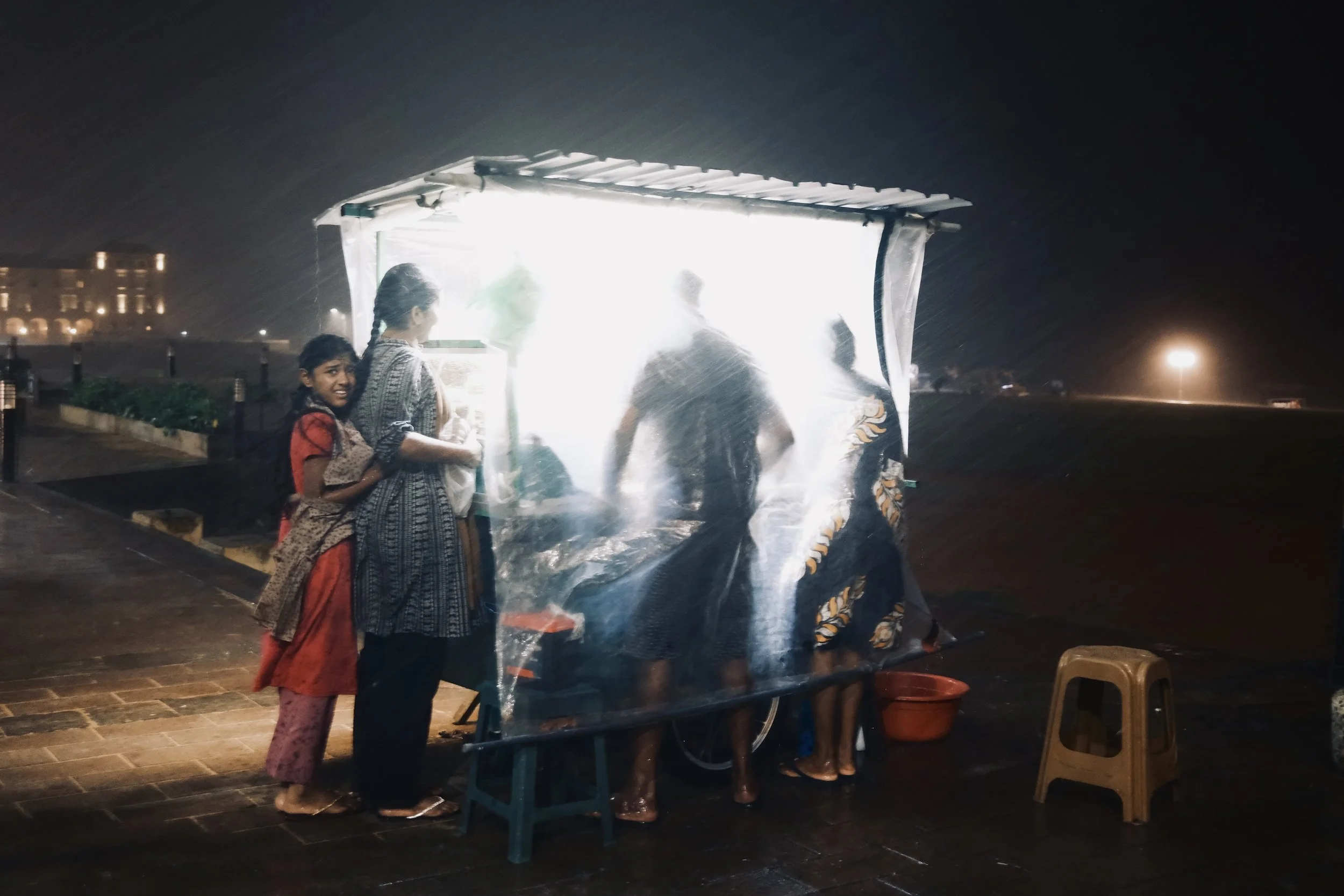 Children and adults standing in line at a small outdoor street food stall during a rainstorm at night, with a bright light illuminating the stall.