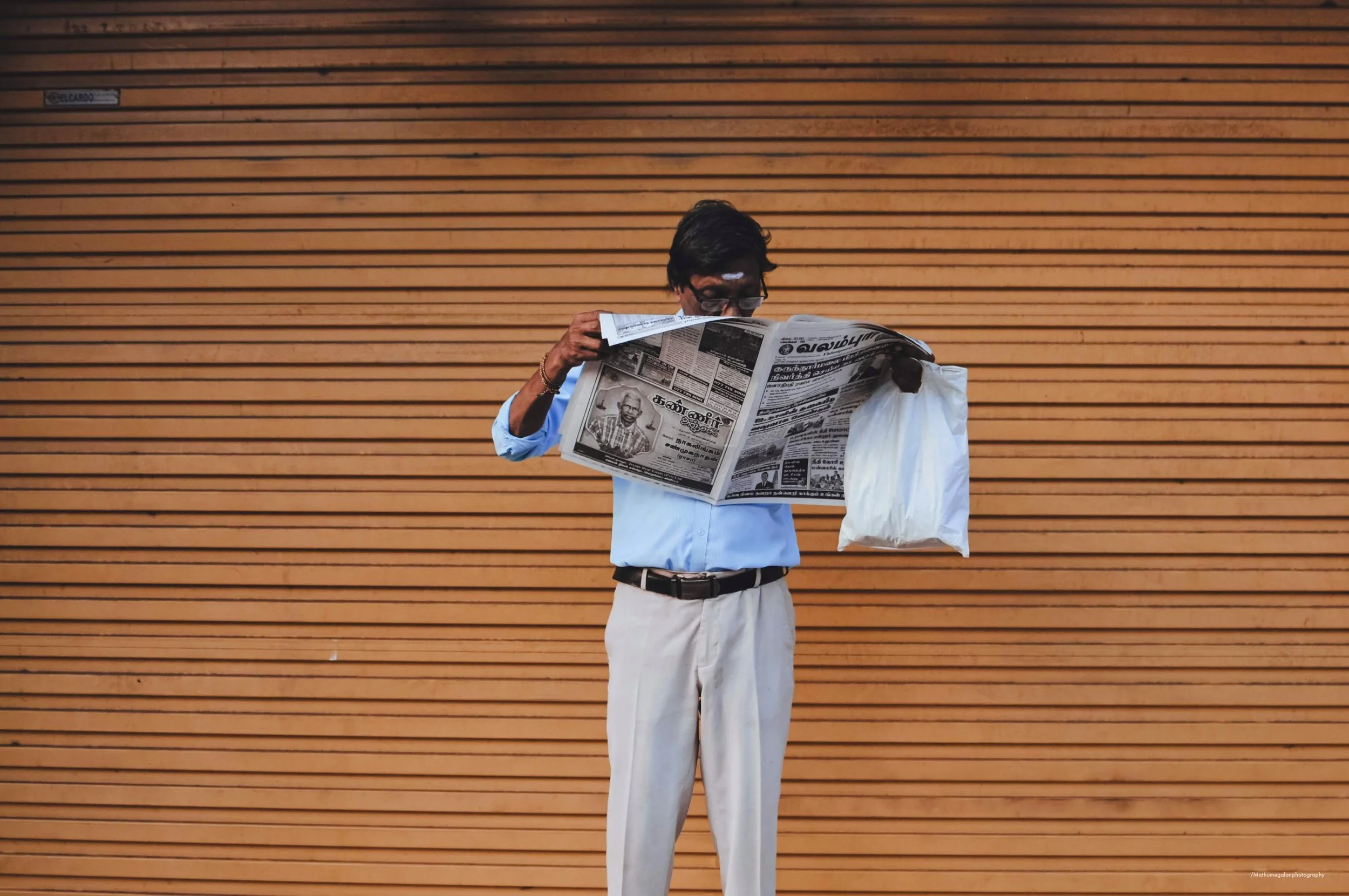 A man with black hair, wearing glasses, a light blue shirt, and beige pants, is reading a newspaper while standing in front of a wooden wall. He is carrying a white bag in his left hand.