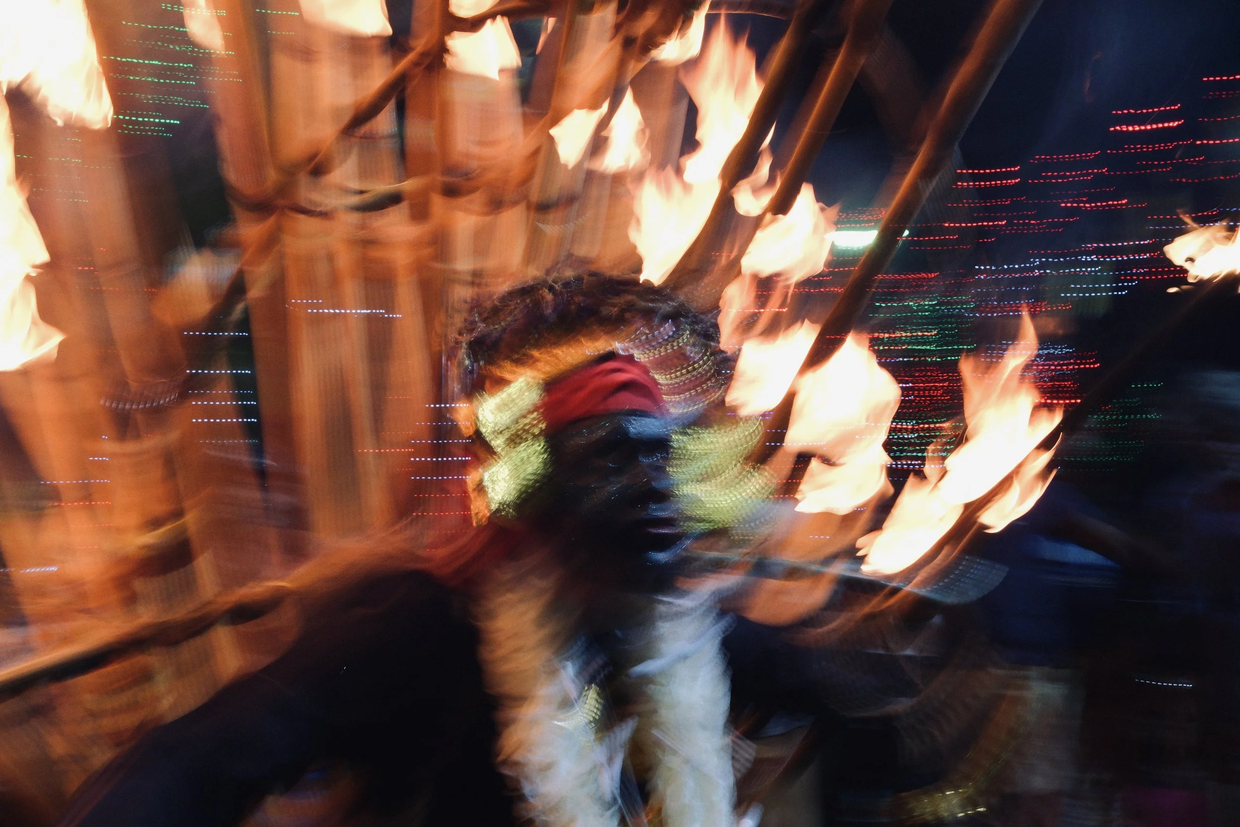 A person at a nighttime event holding a flaming torch, with flames and firelight illuminating their face, surrounded by streaks of colorful lights and blurred motion.