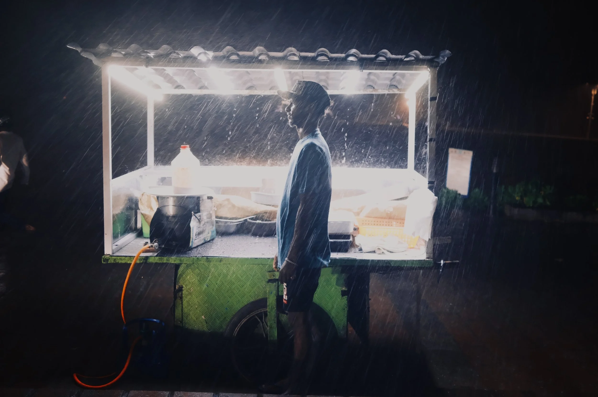 A person with a hat and T-shirt working at a well-lit street food cart during heavy rain at night.