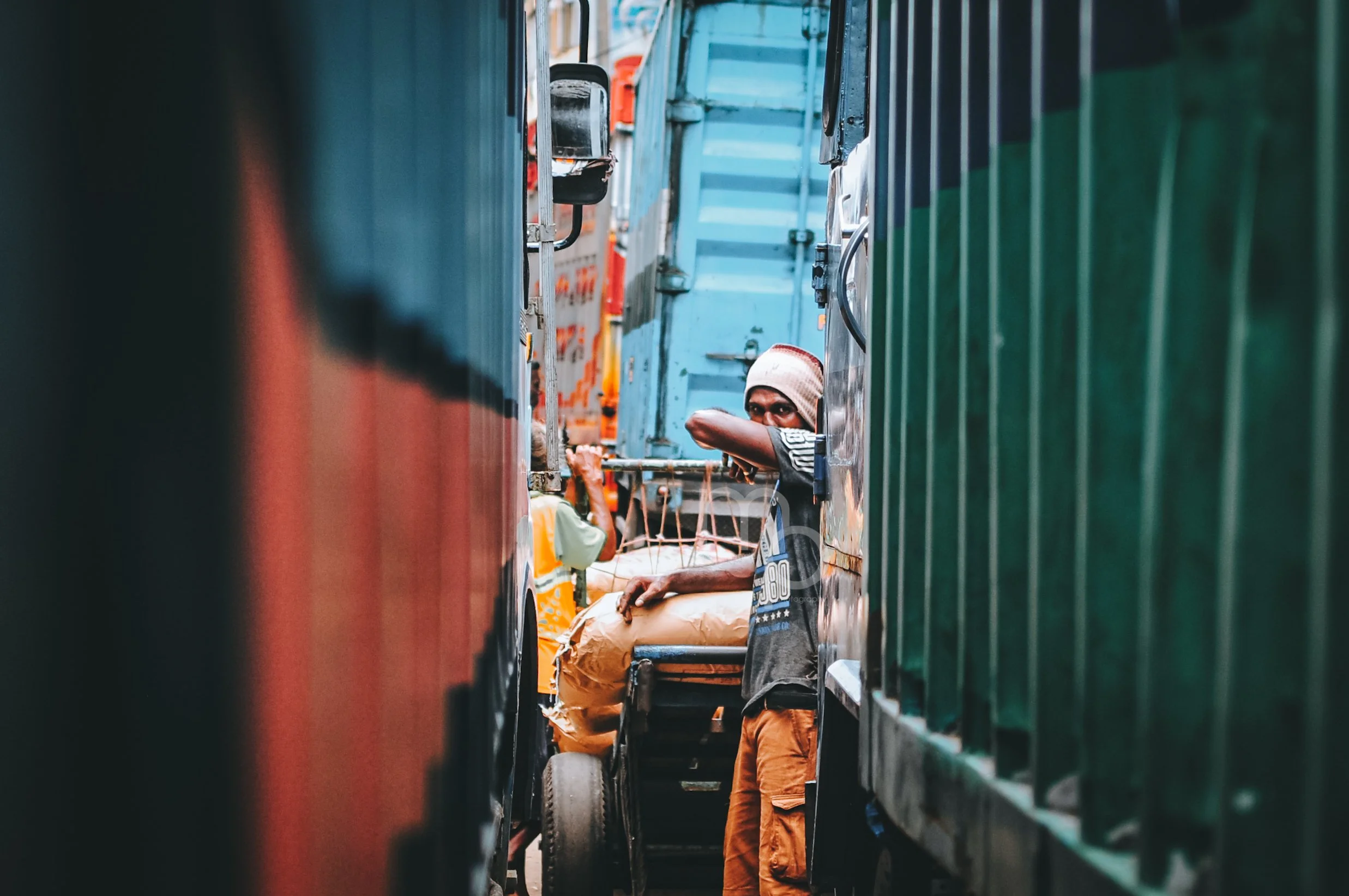 A man wearing a hat and sunglasses looking through a gap between two trucks on a busy street.