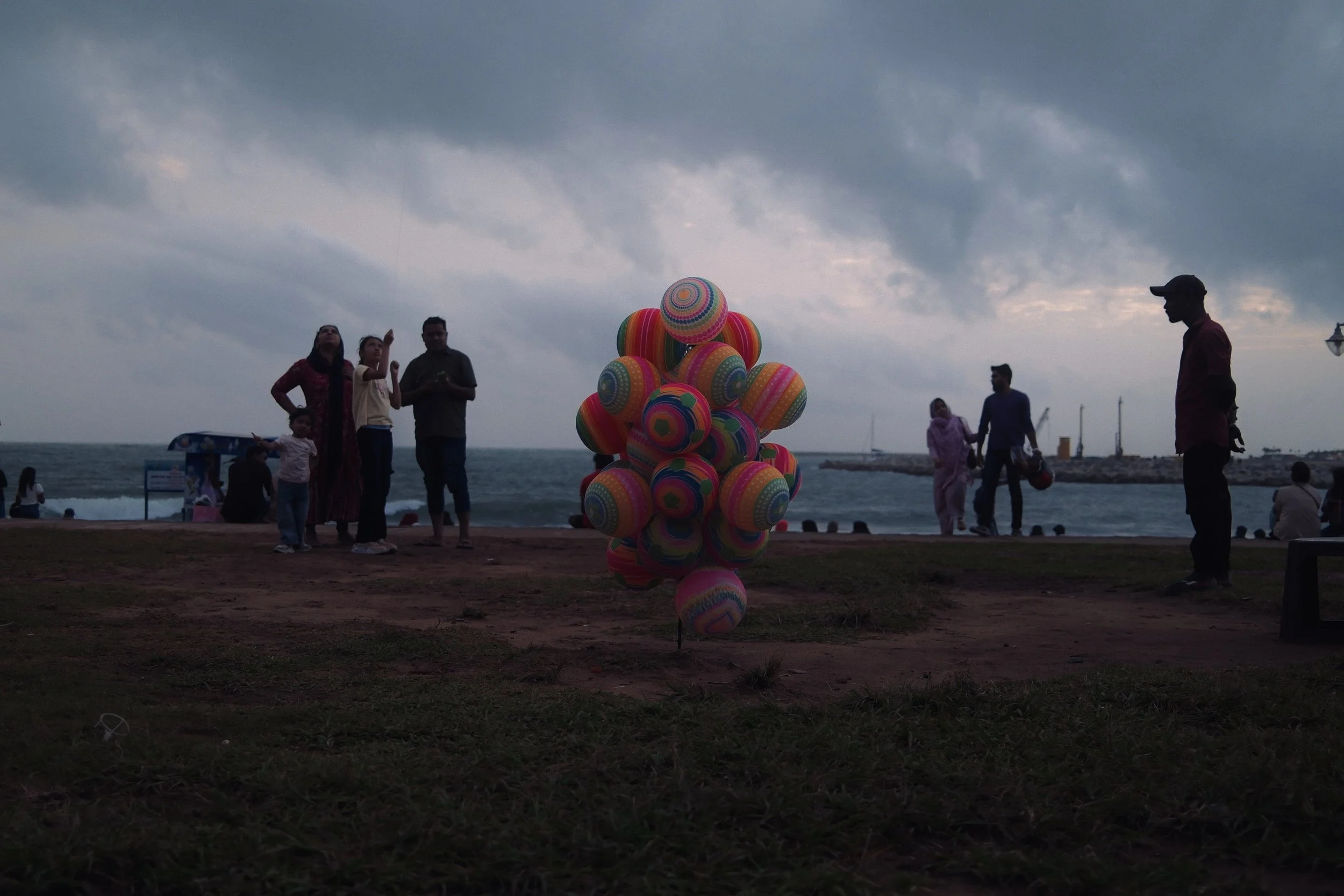 Colorful balloons arranged in a cluster on a beachside promenade with people walking and enjoying the evening near the ocean under a cloudy sky.