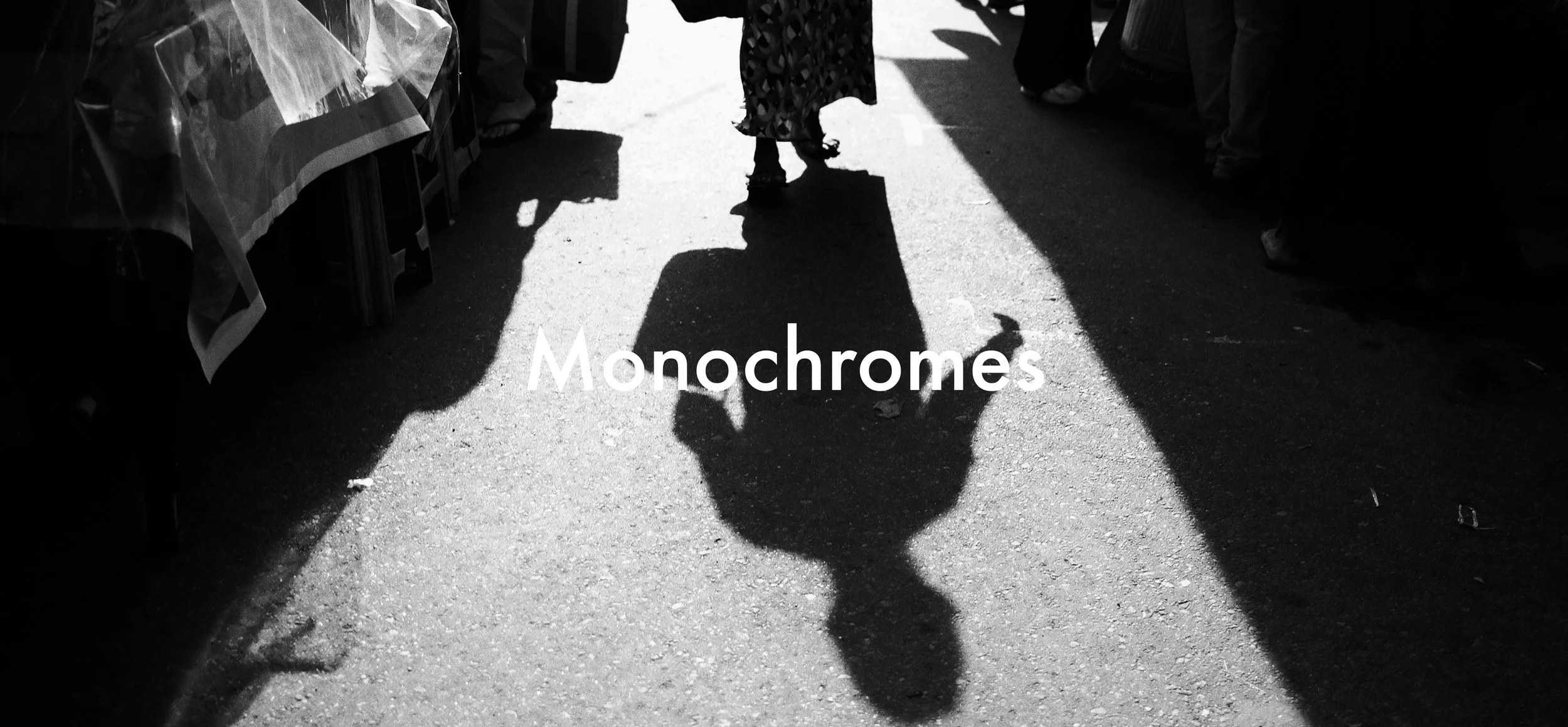 A black and white photo of a person walking at an outdoor market, casting a large shadow on the pavement. The person is wearing a patterned dress or skirt. The word "Monochromes" is overlaid on the image.