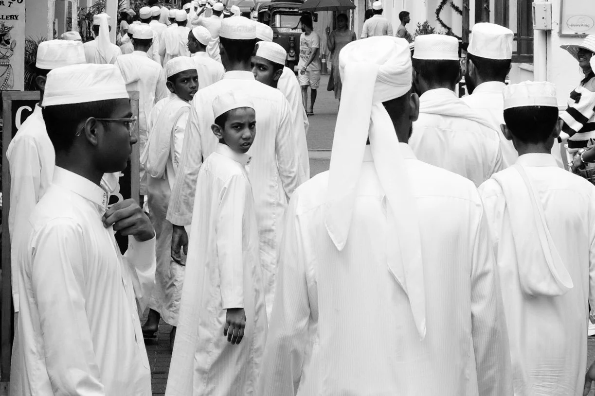 Group of young boys dressed in white traditional clothing and caps, walking down a street, with some looking back and others ahead, among shops and passersby.