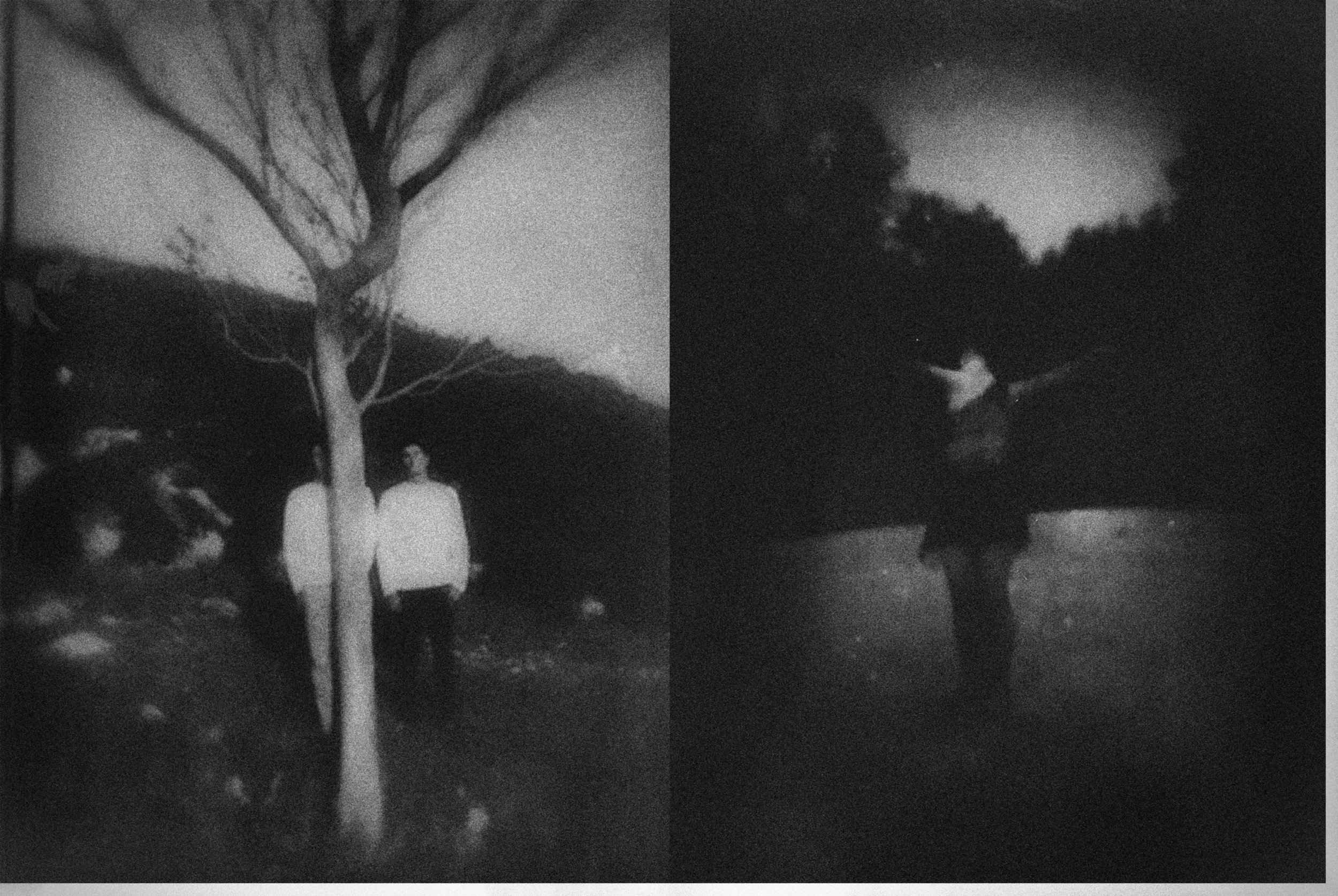 Black and white photo of two people standing outdoors at night with trees and cloudy sky in the background. The person on the left is wearing a light-colored hoodie, and the person on the right is wearing a dark jacket with a hoodie. The image appear