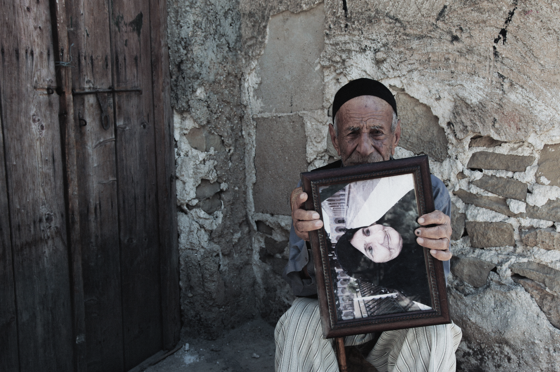 An elderly man with a black cap is holding a framed photo of a young girl and children. He is seated against a rustic stone wall, creating a poignant scene.