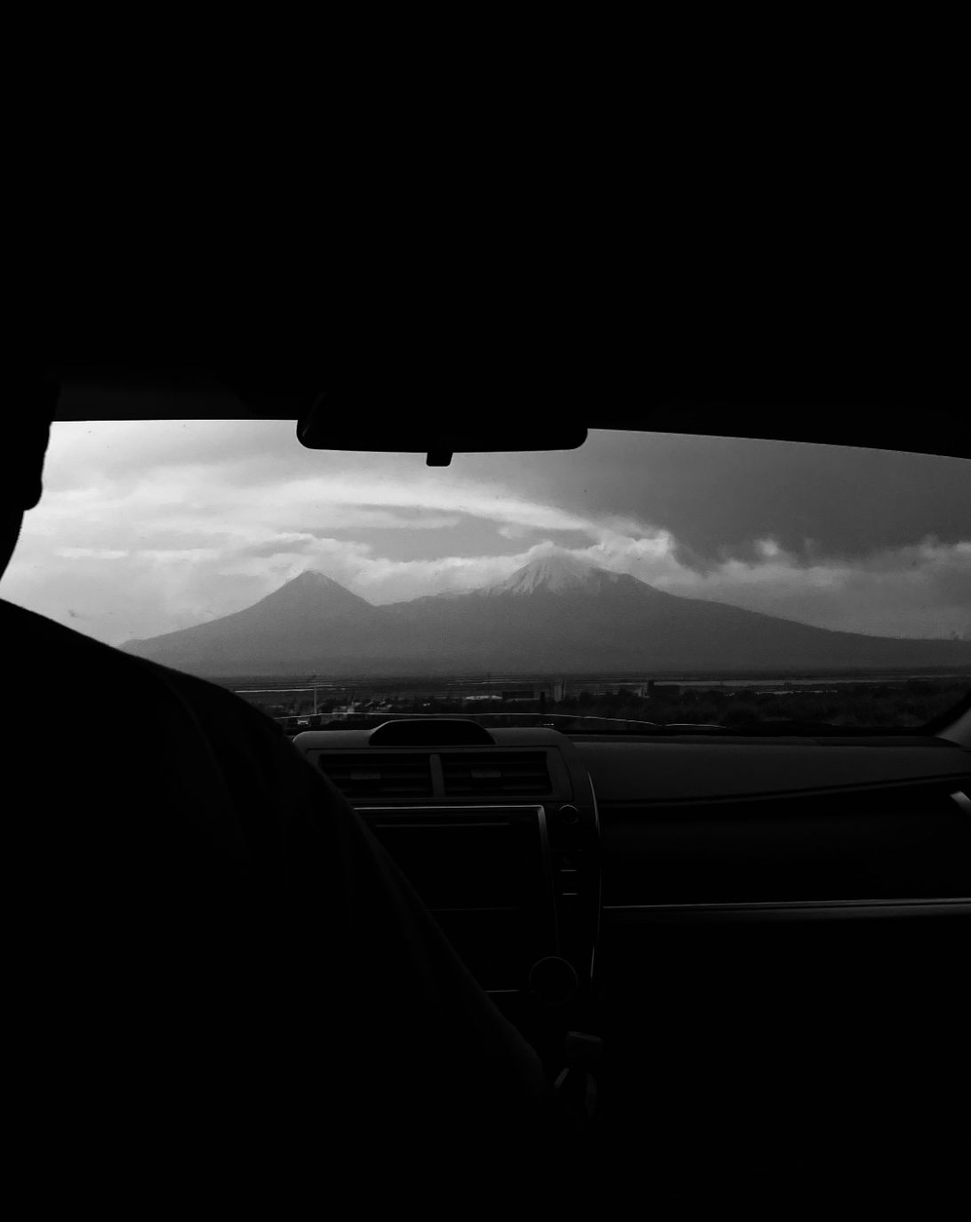 Black and white photo of two people in a car, with a view of a mountain range, including Mount Fuji, through the windshield.