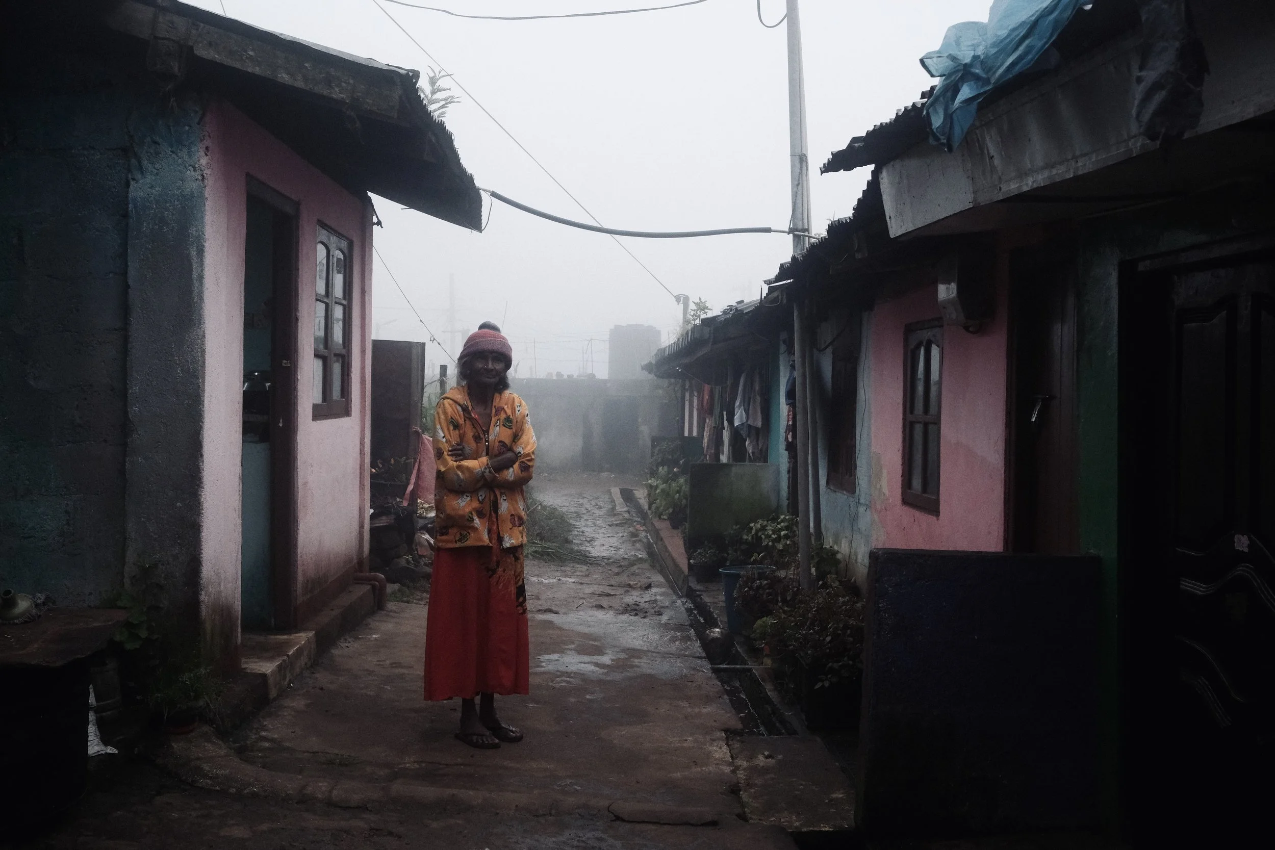 A woman standing on a muddy street between small, colorful houses in a foggy, rural area.
