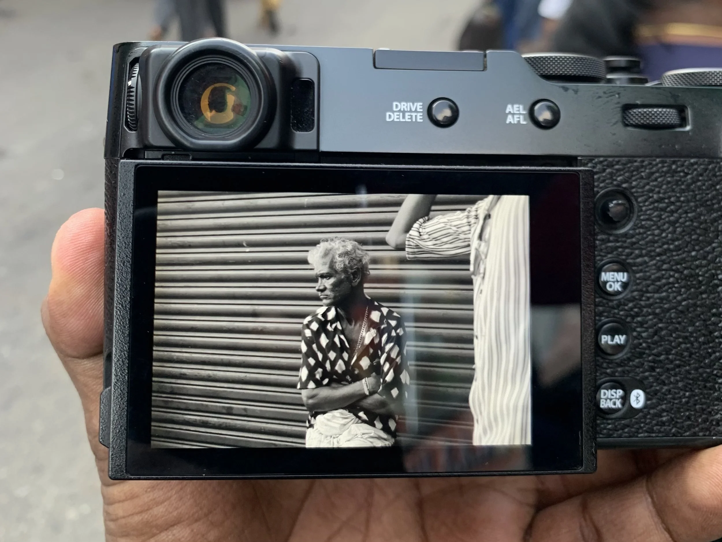 A person holding a camera capturing an image of an elderly woman with gray hair, wearing a patterned shirt, standing against a metallic corrugated wall.