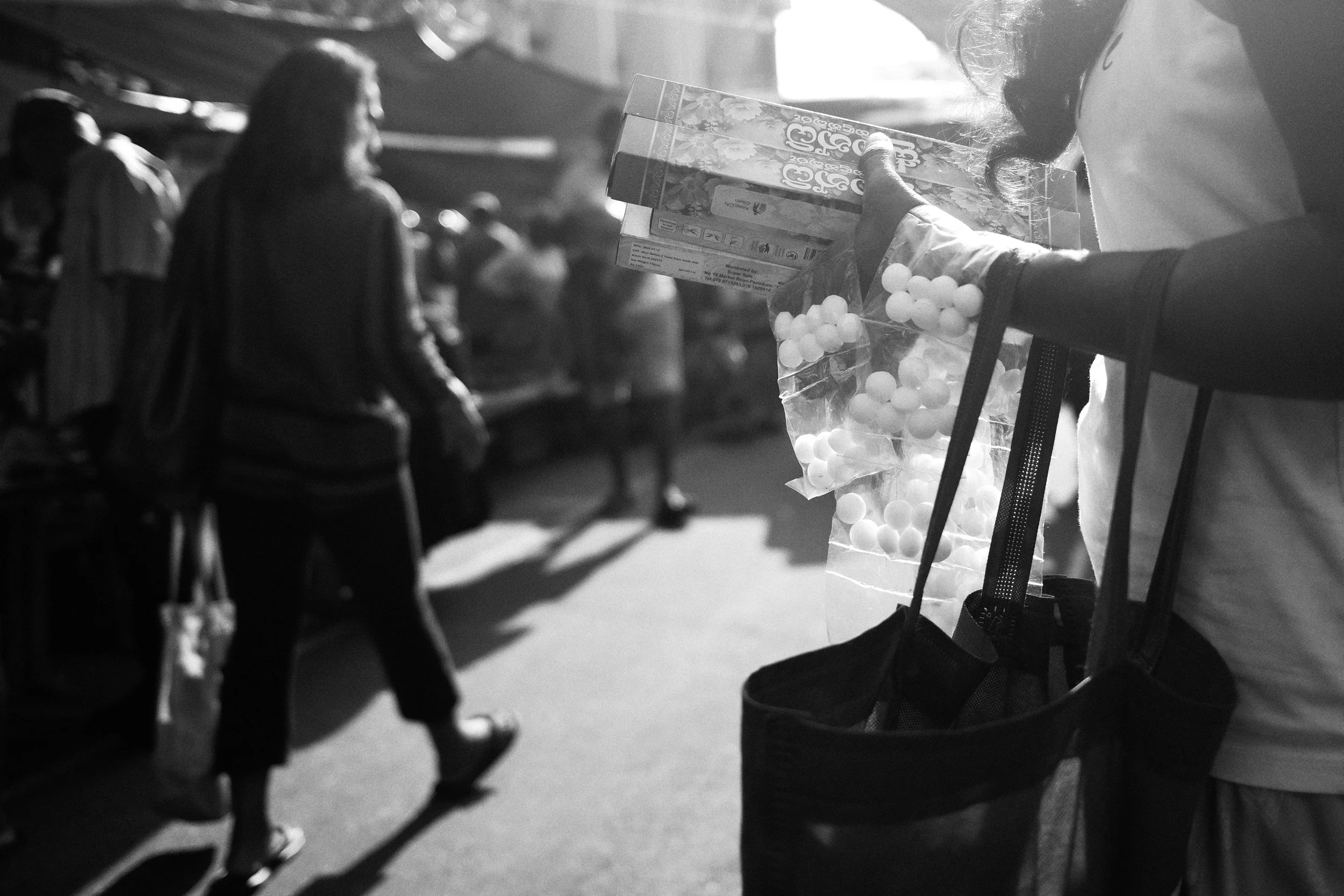 A street vendor holding a bag of small round balls and a box of candy in a busy outdoor market with people walking by.